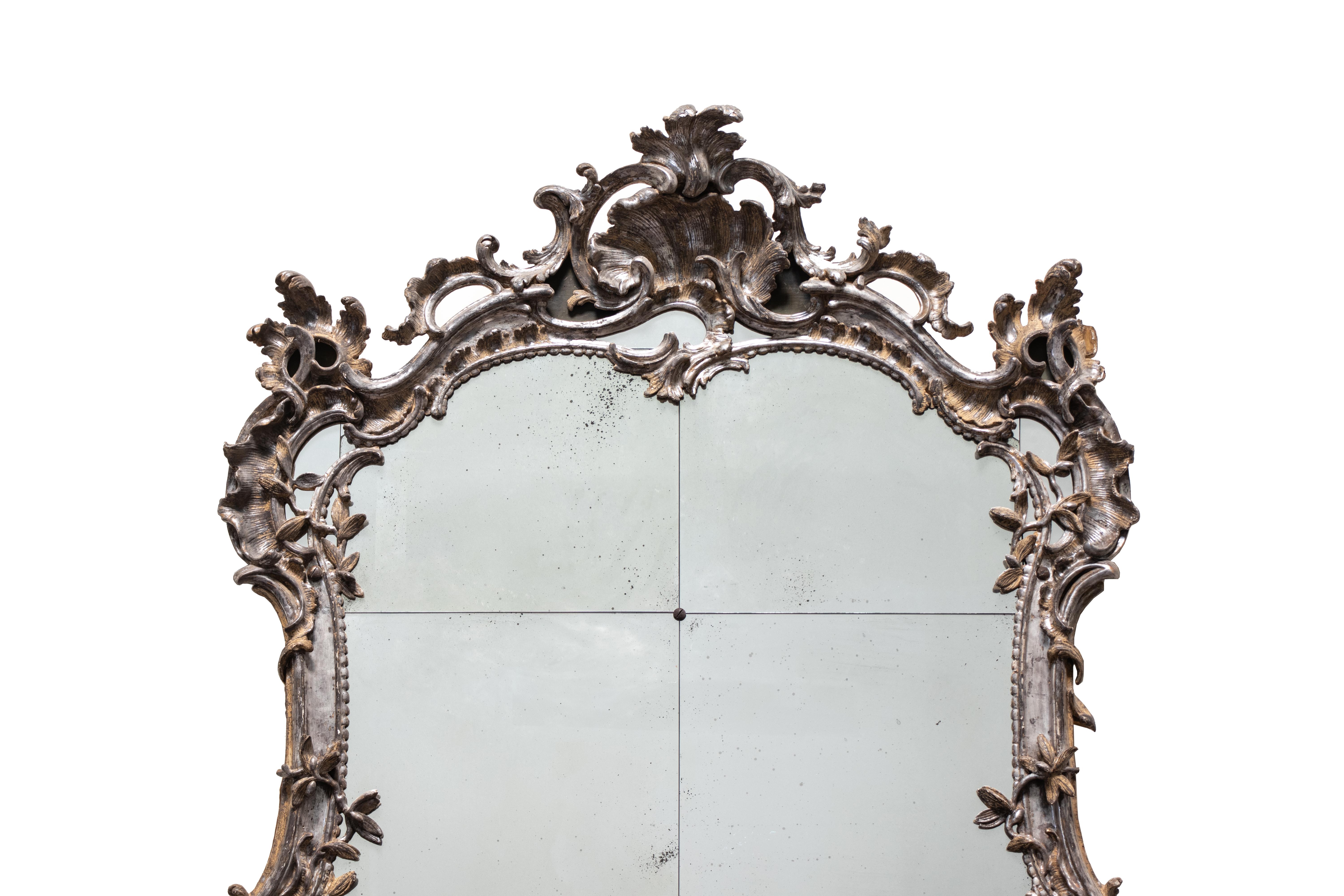 A grand-scale wall or console mirror in Rococo style from mid-18th century South Germany. It features six mirror panels framed in richly decorated silvered wood, carved in the shapes of rocaille scrolls and foliage, with two dragon figures at the