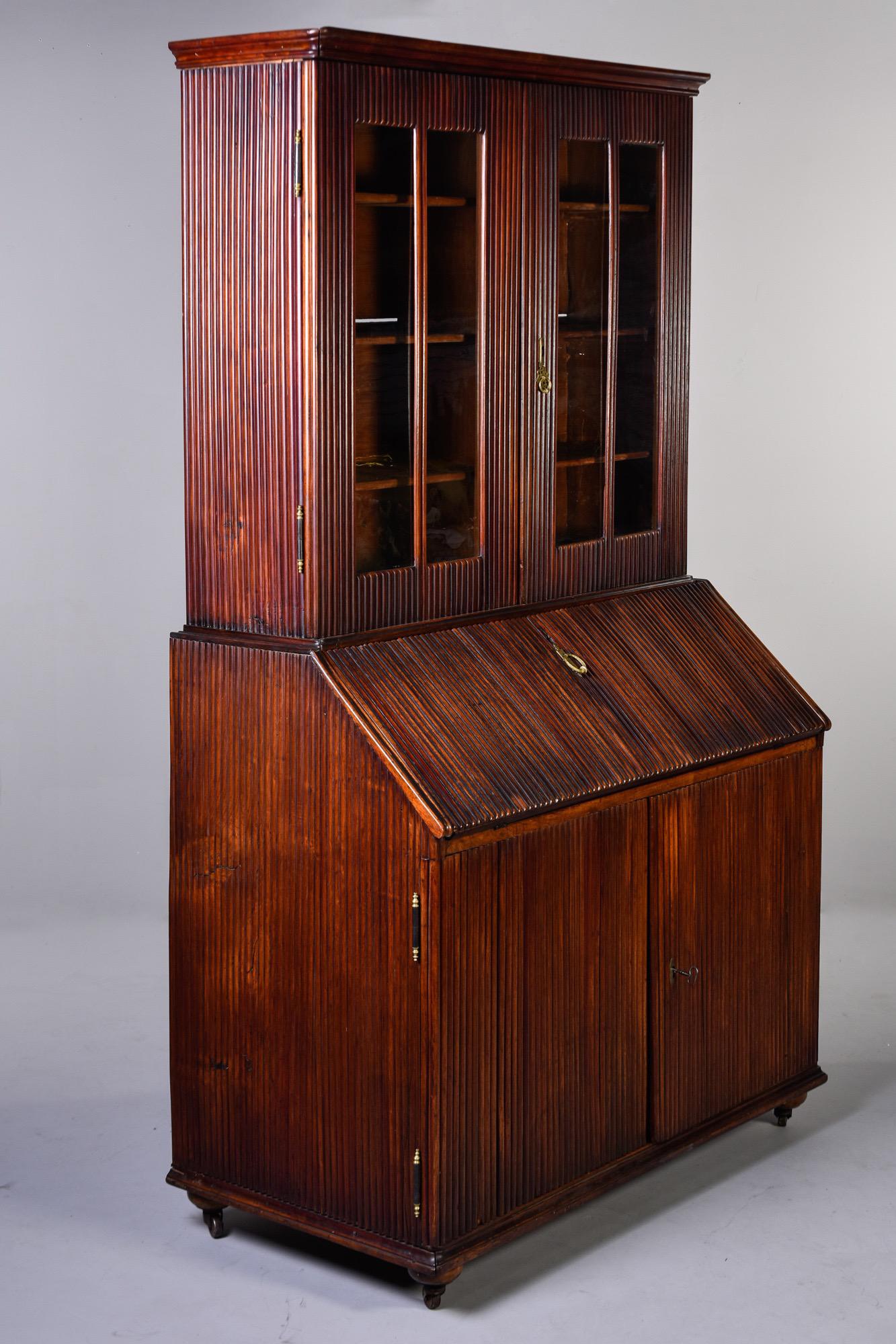 English cabinet features a reeded walnut base with fold down desk, internal shelves at the bottom, caster feet and glass-fronted book shelves on the top, circa 1850s. Unknown maker. Very good overall antique condition with crack on front of base and