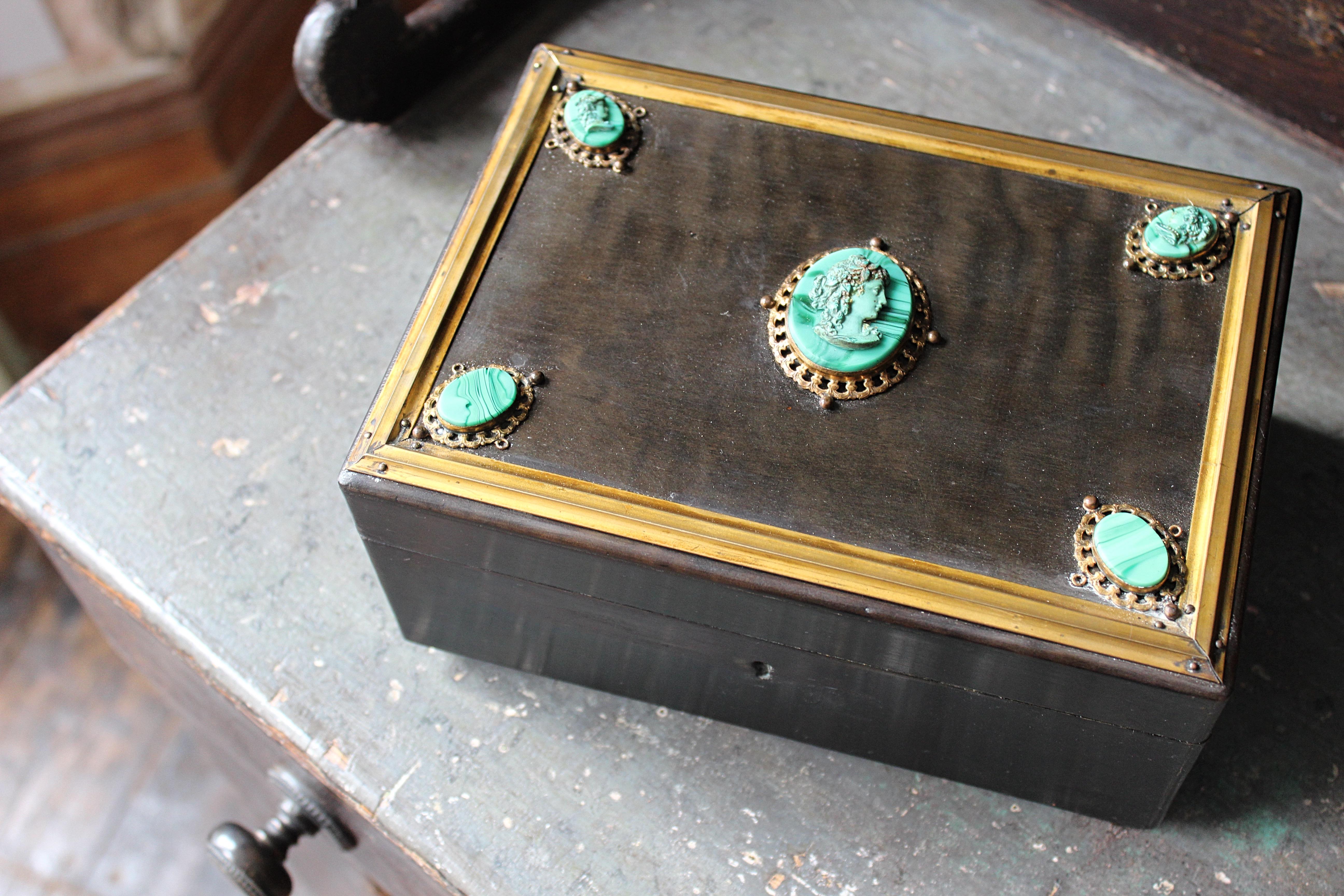 A good quality brass bound and ebonised trinket/jewellery box, decorative with malachite stones and cameos.

The interior is a lovely faded blue silk with decorative pipping, with its original cast brass hinges and a wooden caucus

Age related