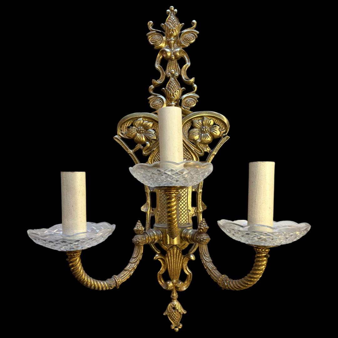 Mid 19th-century brass 3 arm antique wall lights.

Crafted with exquisite twisted arms and adorned with a hand-chased floral design on the back plate, these fixtures exude vintage charm.
The bobeche glass drip trays add an extra touch of elegance