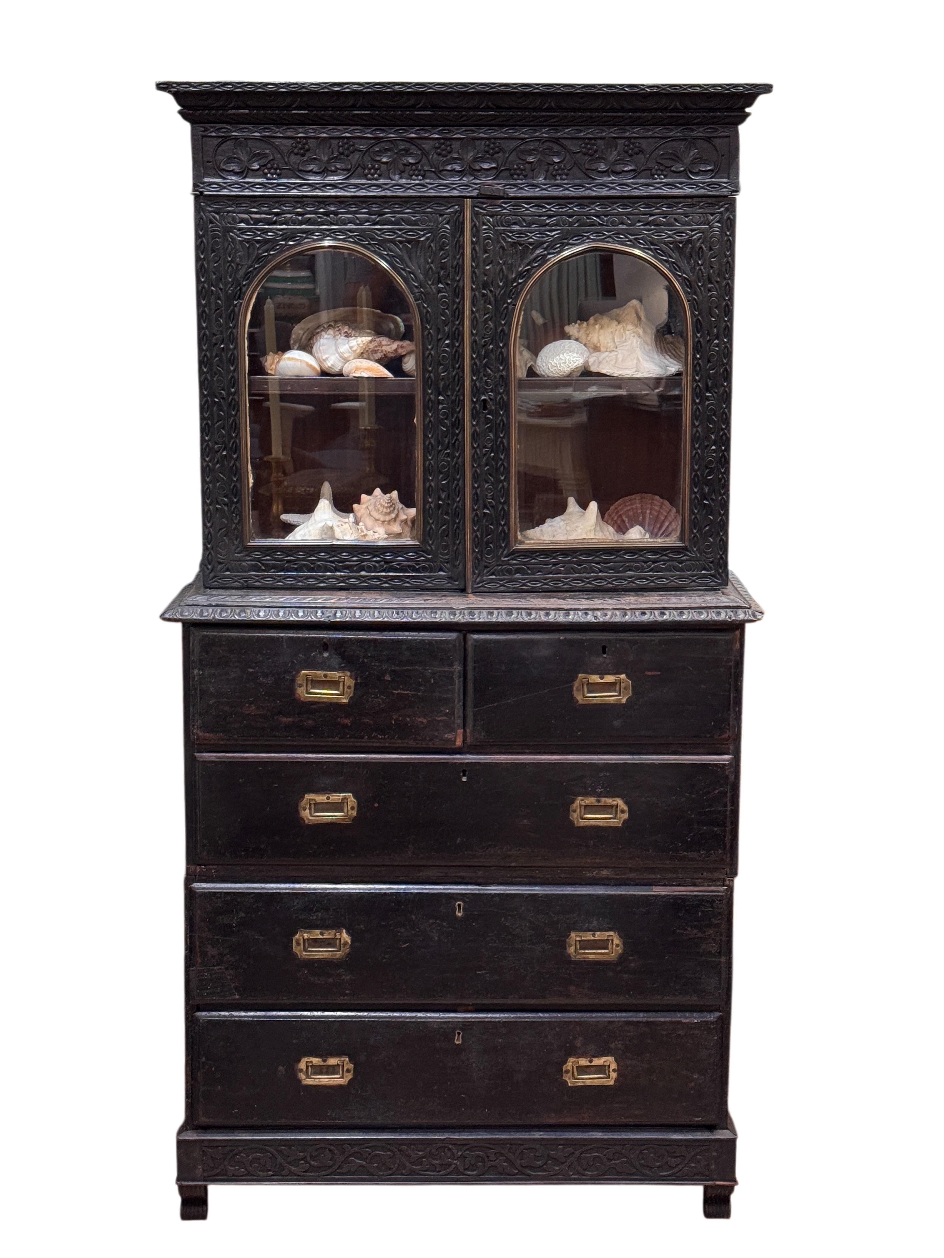 Arcón de campaña angloindio de ébano tallado, muy poco común, en tres secciones, hacia 1850. El mueble superior presenta motivos foliados y enredaderas profundamente tallados que enmarcan un par de puertas acristaladas arqueadas, sobre cuatro
