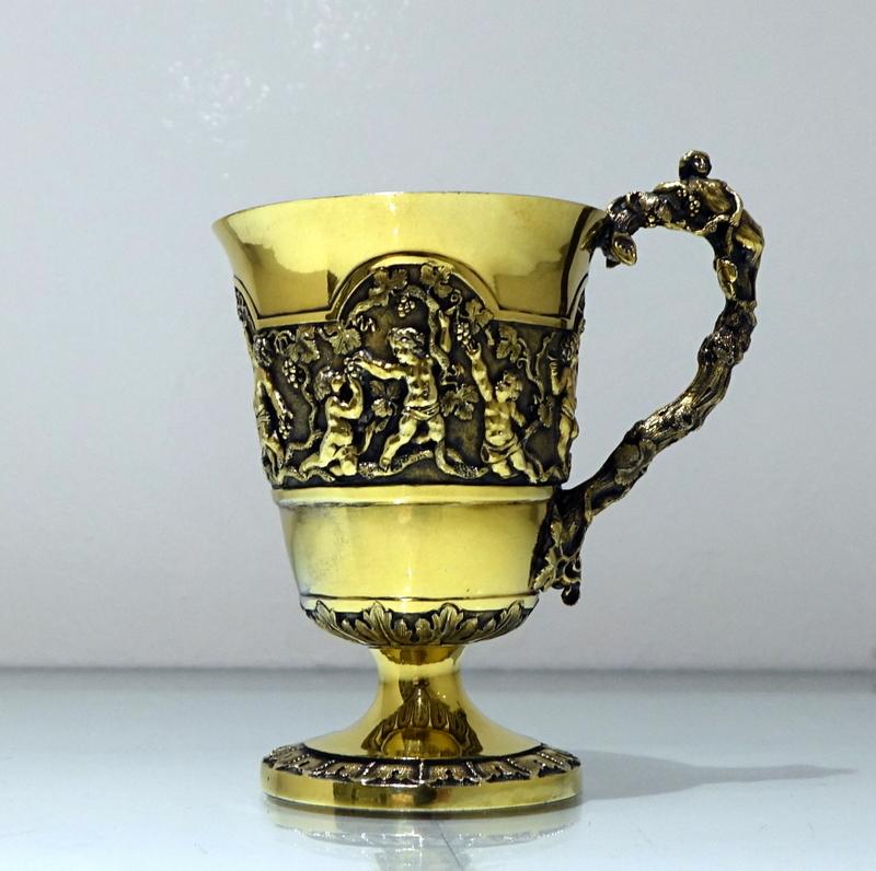 A magnificent mid-19th century silver gilt christening mug decorated with beautiful scenes of cherubs picking grapes from a climbing vine shrub which is then set on a matte background for decorative contrast. The mug has a stunning cast naturalistic