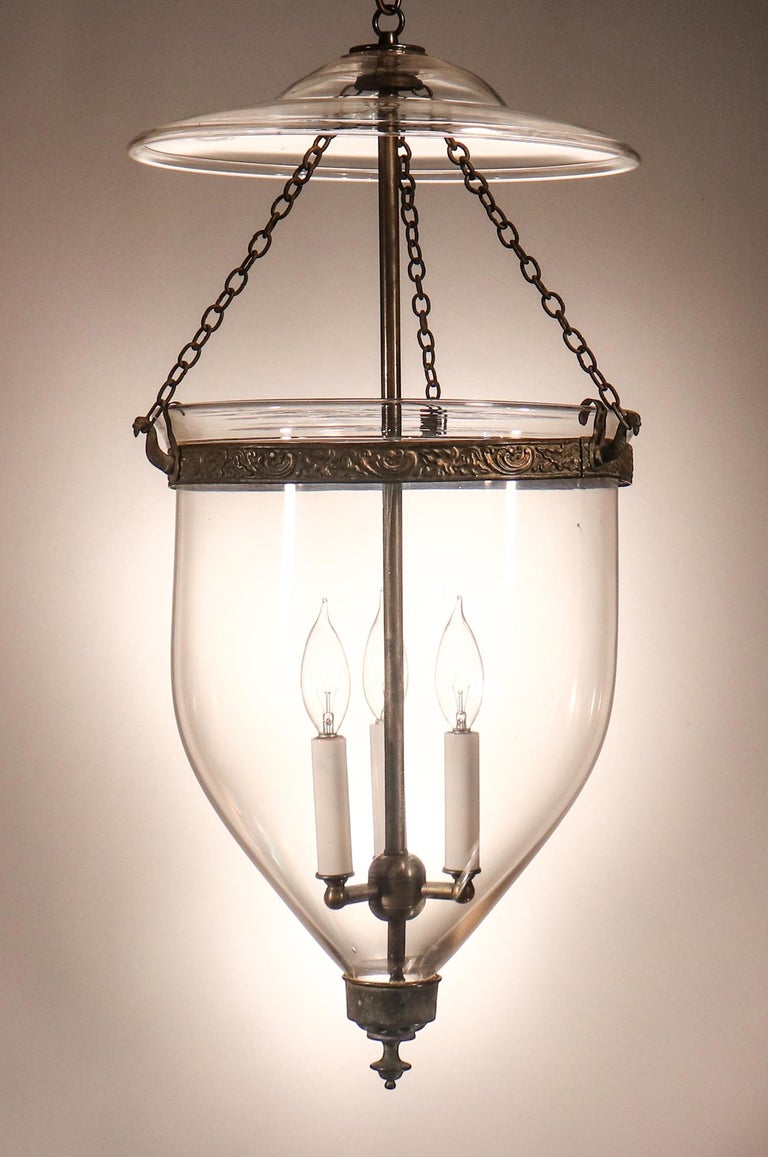 An antique English bell jar lantern with elegant form and authentic brass fittings—including a rare embossed band with tassel motif that adds just the right amount of ornamentation to this hand blown clear glass lantern. The circa 1850 pendant has