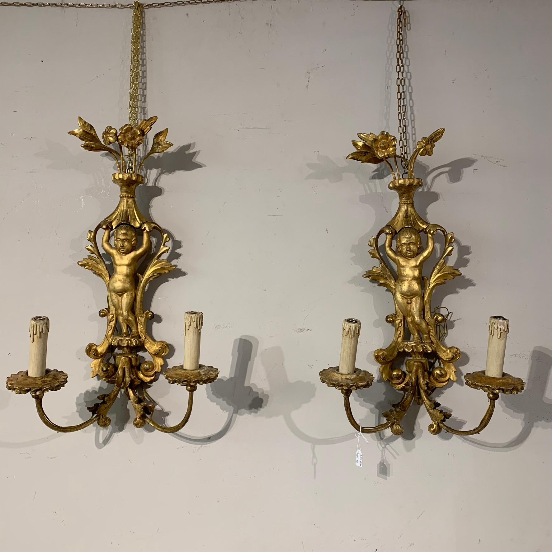 Beautiful pair of carved and gilded wooden appliques with pure gold leaf, made like two cherubs holding up a vase of flowers.
Two candle holder arms in iron, also gilded.