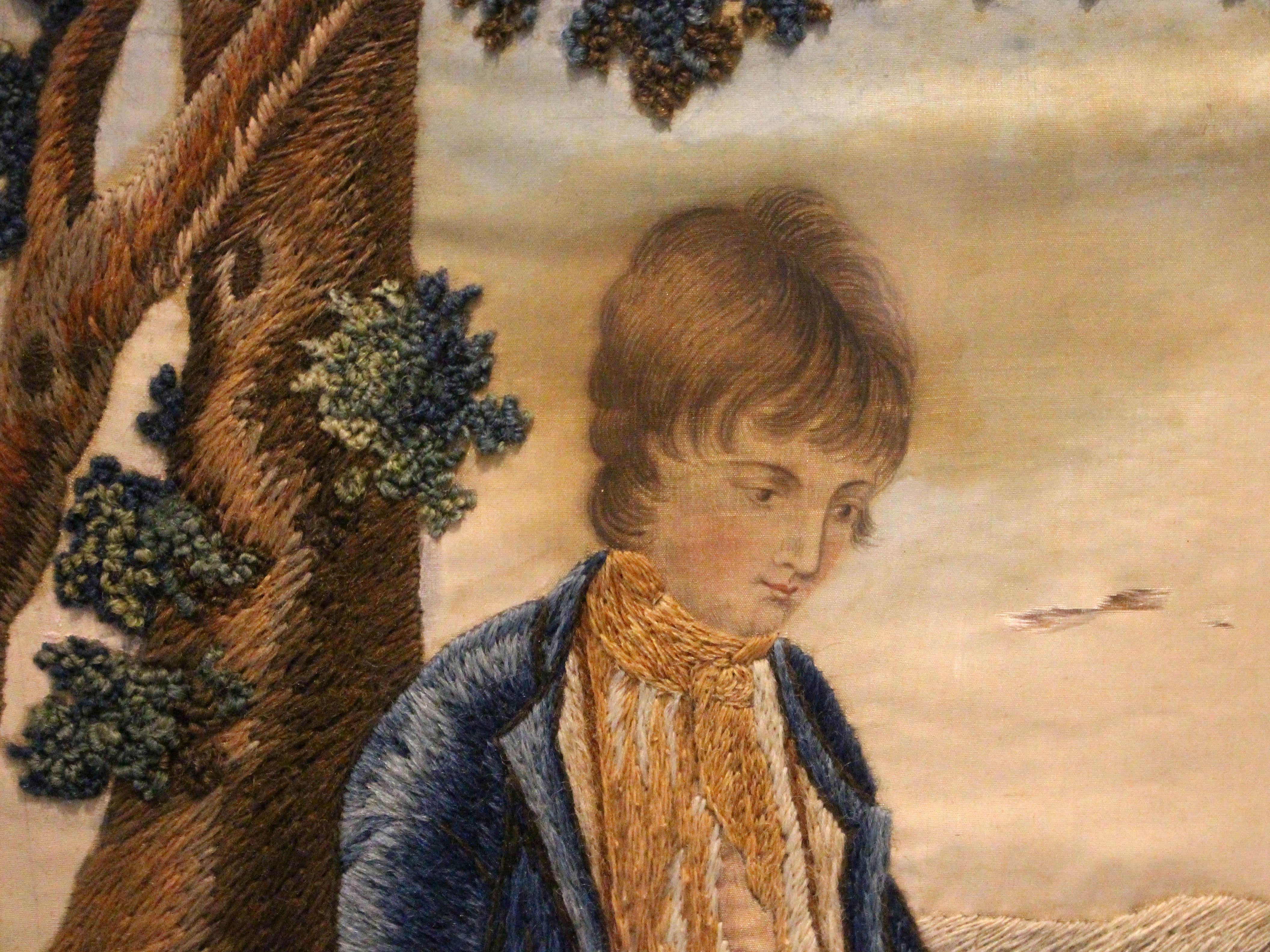 Mid-19th century embroidery work on silk, framed, English. A charming scene of a lad leaning by a tree with his dog hopefully awaiting a treat of the bread the boy is cutting; by a gate to the rolling English countryside. The silk with some
