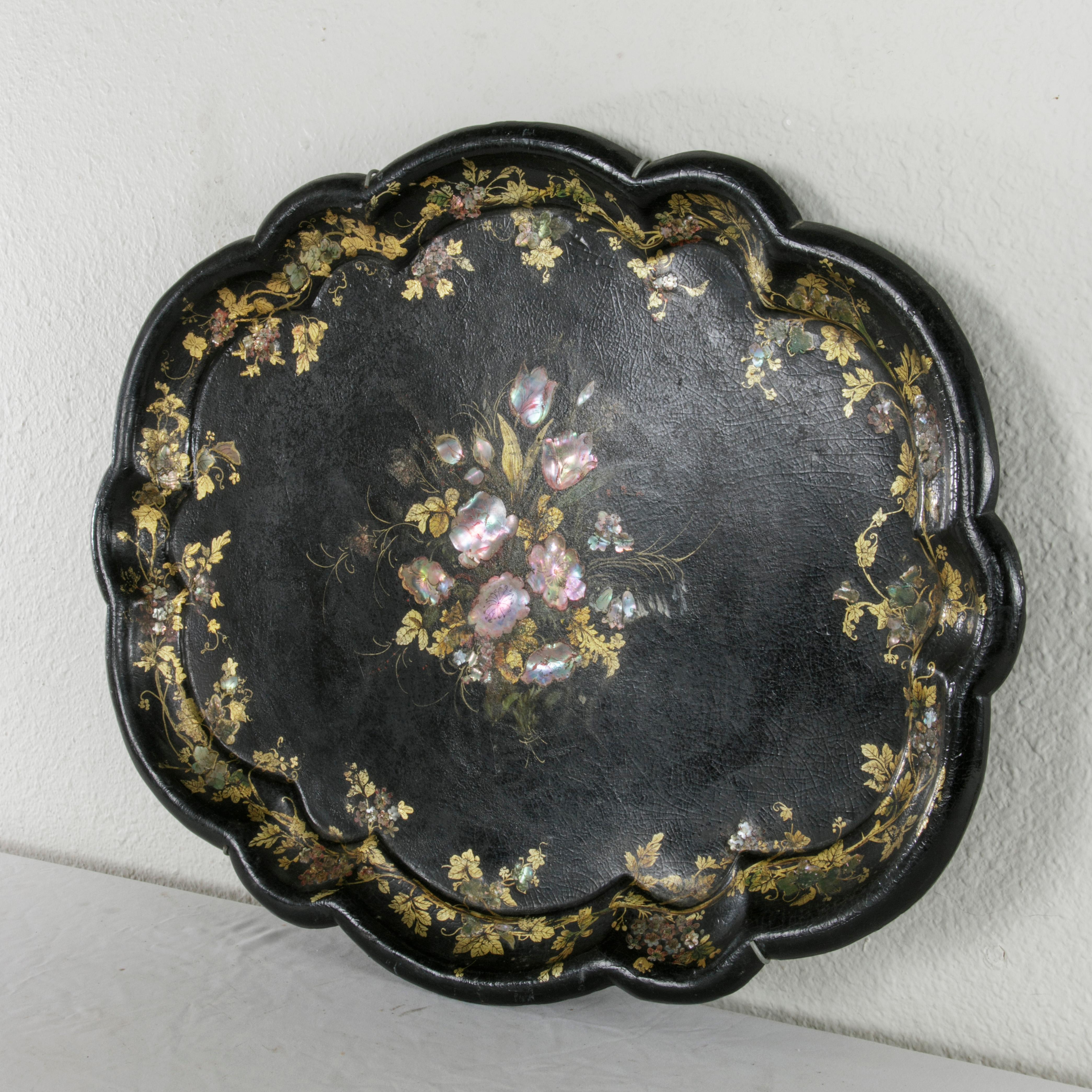 This mid-19th century English Victorian period black papier mâché tray features a central inlaid mother-of-pearl flower arrangement with hand-painted gold detailing of a variety of leaves. The floral and leaf motif is repeated around the scalloped