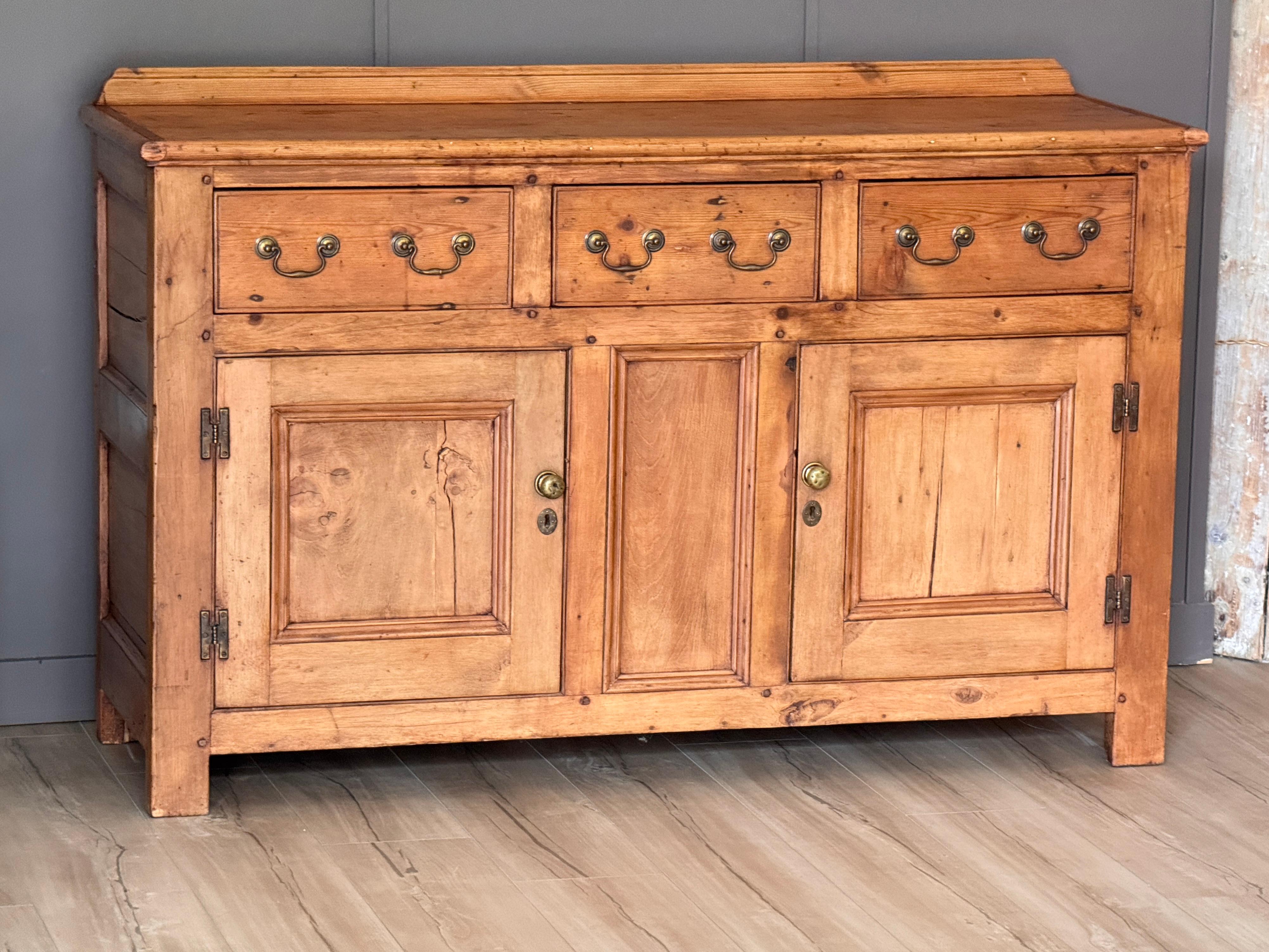 Cabinet anglais du milieu du C.I.C. en pin ciré à la finition naturelle et chaleureuse. La pièce comporte trois tiroirs sur des portes à panneaux avec des poignées et des écussons en laiton. Les lignes droites et la construction épurée soulignent