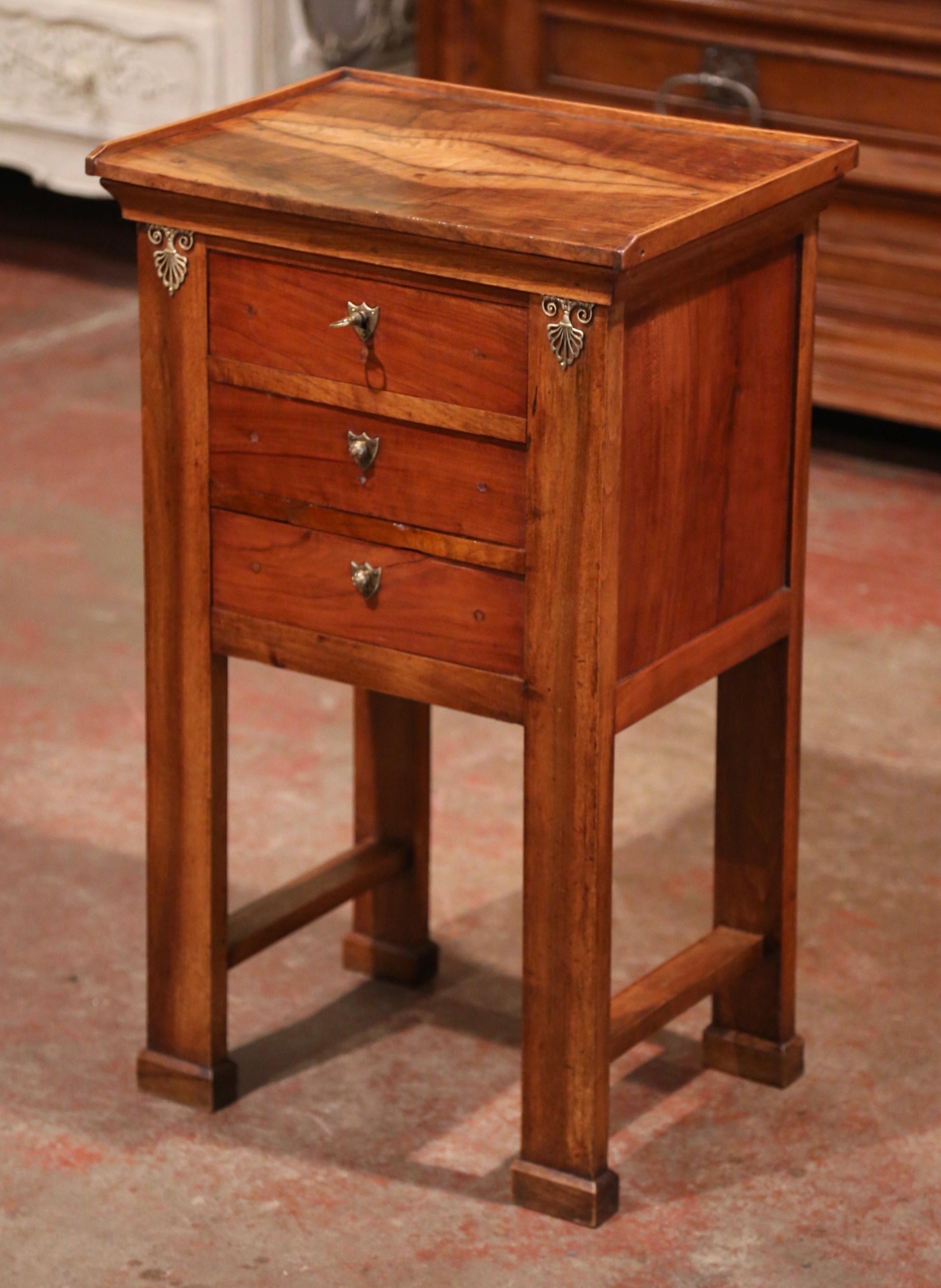 This elegant antique nightstand was crafted in France, circa 1850. The traditional fruit wood cabinet stands on four straight legs terminated with plinth feet and embellished with decorative brass mounts; the small chest features three drawers