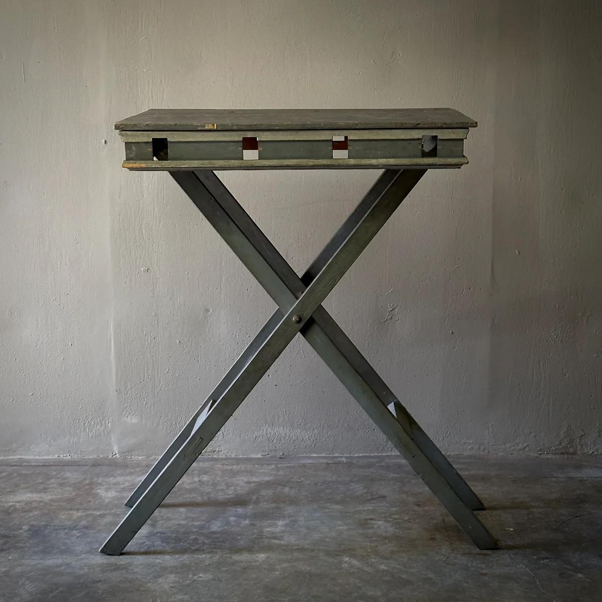 This presentation table boasts an industrial yet delicate appeal with its foldable X-shaped metal base and gray painted wooden top. Originally used for displaying models or sculptures, it features a worn surface that narrates its history through