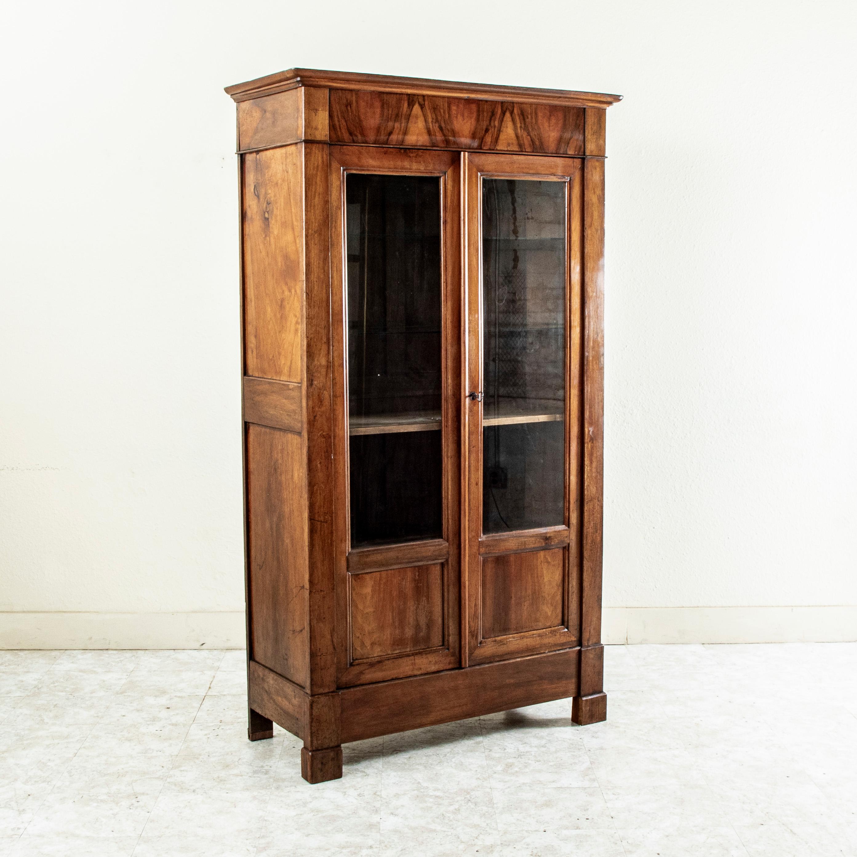 Questa libreria o vetrina francese d'epoca Louis Philippe del XIX secolo presenta una facciata in noce massiccio dalle linee semplici e belle e due ante incastonate con vetro. Un pannello sulla corona è decorato con un motivo abbinato al libro.