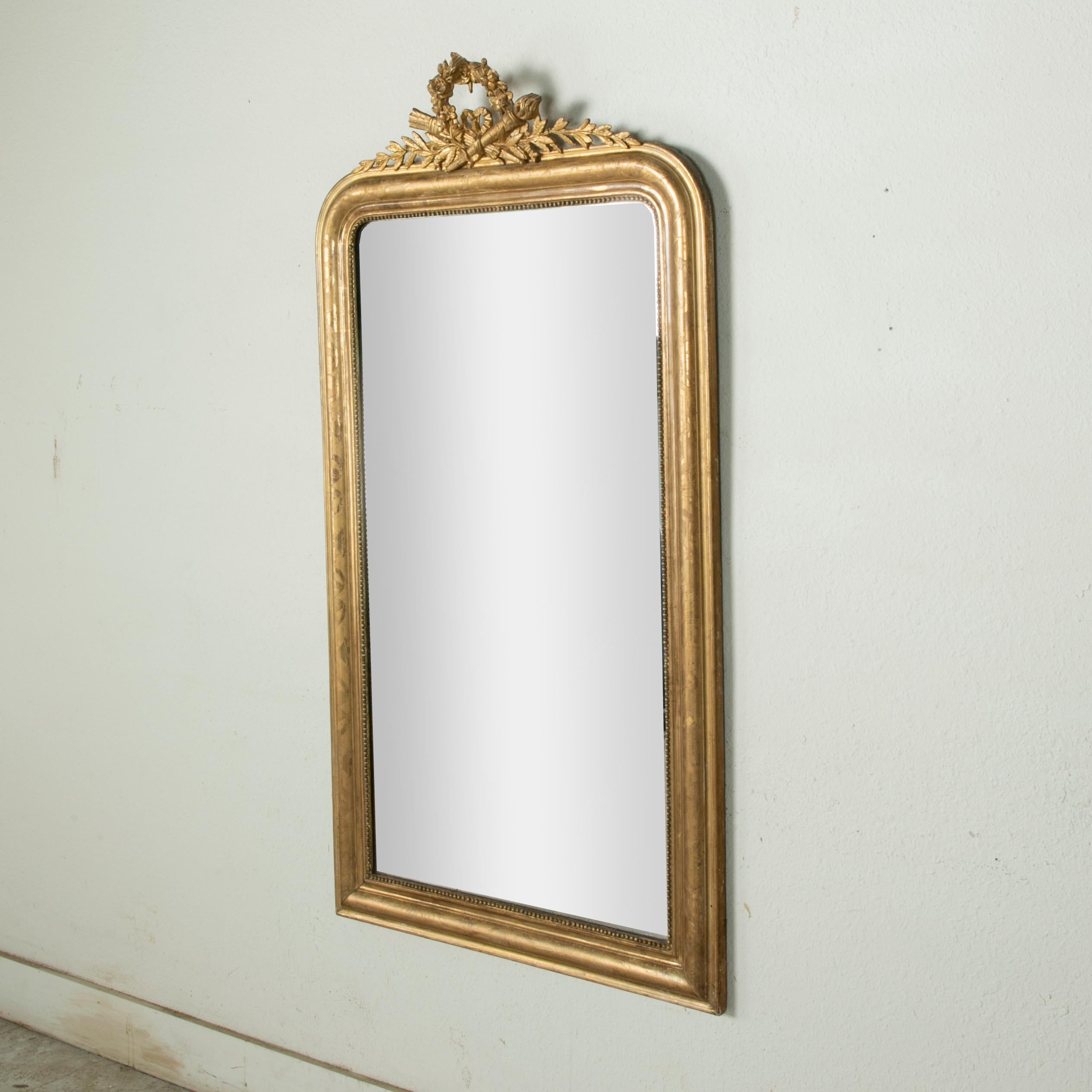 This mid-nineteenth century Louis Philippe style gilt wood mirror features its original mercury glass surrounded by a pillow frame detailed with subtle incising in a floral motif and an inner border of fine beading. A classic Louis XVI crossed torch