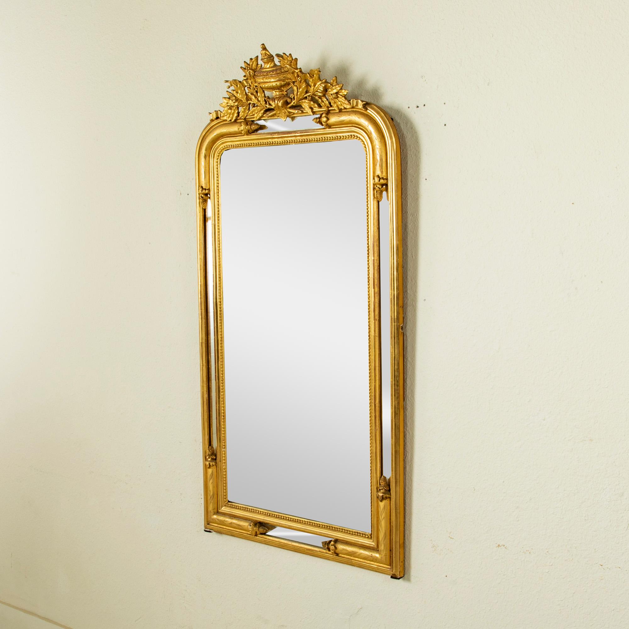 Ideal above a mantel, buffet or chest of drawers, this elegant mid-nineteenth century giltwood Louis XVI style mirror boasts its original beveled mercury glass. Its ornate frame is hand-carved with an urn at the crown flanked by scrolling ribbons