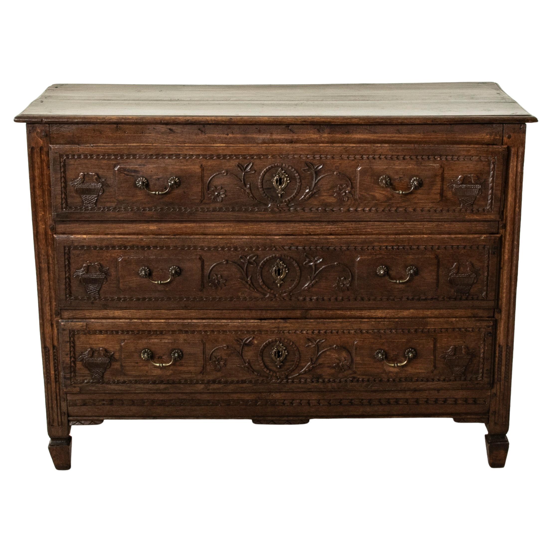 This mid nineteenth century French Louis XVI style oak commode or chest of drawers features hand pegged paneled sides and drawer fronts with hand carvings of scrolling leaves and flowers, and woven baskets with birds. Its three dove tailed drawers