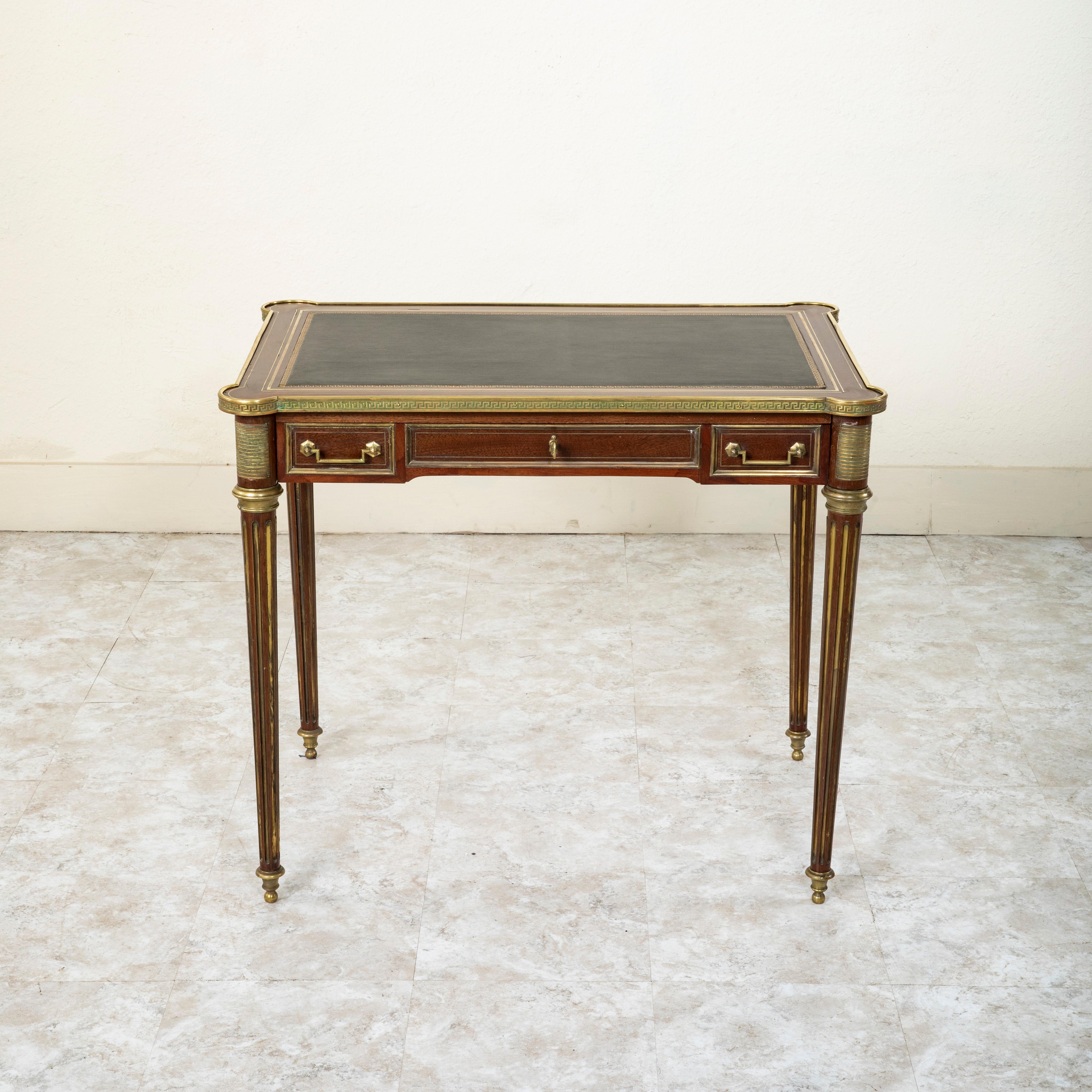 This small scale French Louis XVI style writing table or desk from the mid 19th century is constructed of solid mahogany and features a gold tooled black leather writing surface with a Greek key motif. Two pull out trays are additionally fitted with