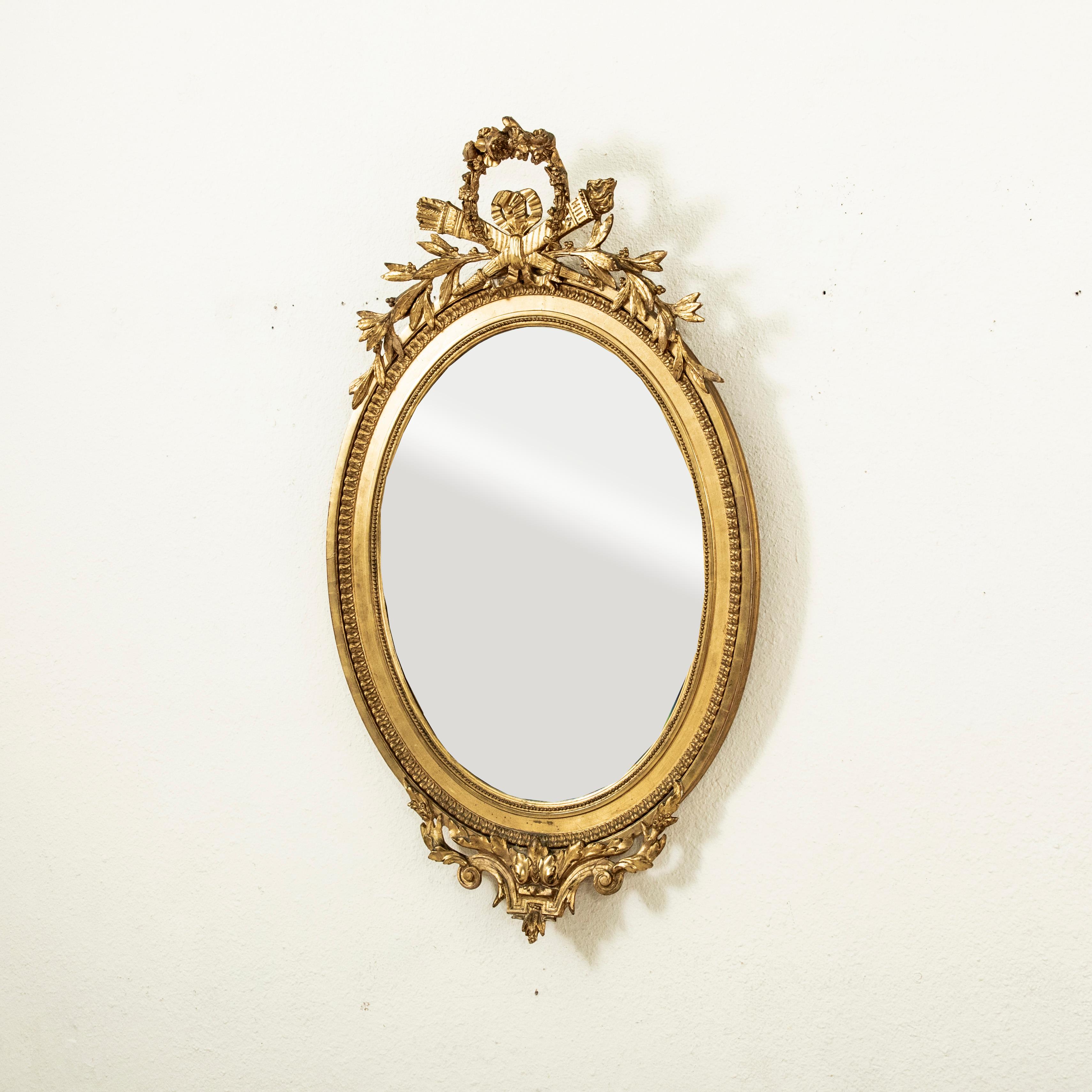 Ce miroir ovale en bois doré de style Louis XVI, datant du milieu du XIXe siècle, est équipé de son verre au mercure biseauté d'origine et présente une couronne de fleurs ainsi qu'une torche et un carquois croisés classiques dans la partie