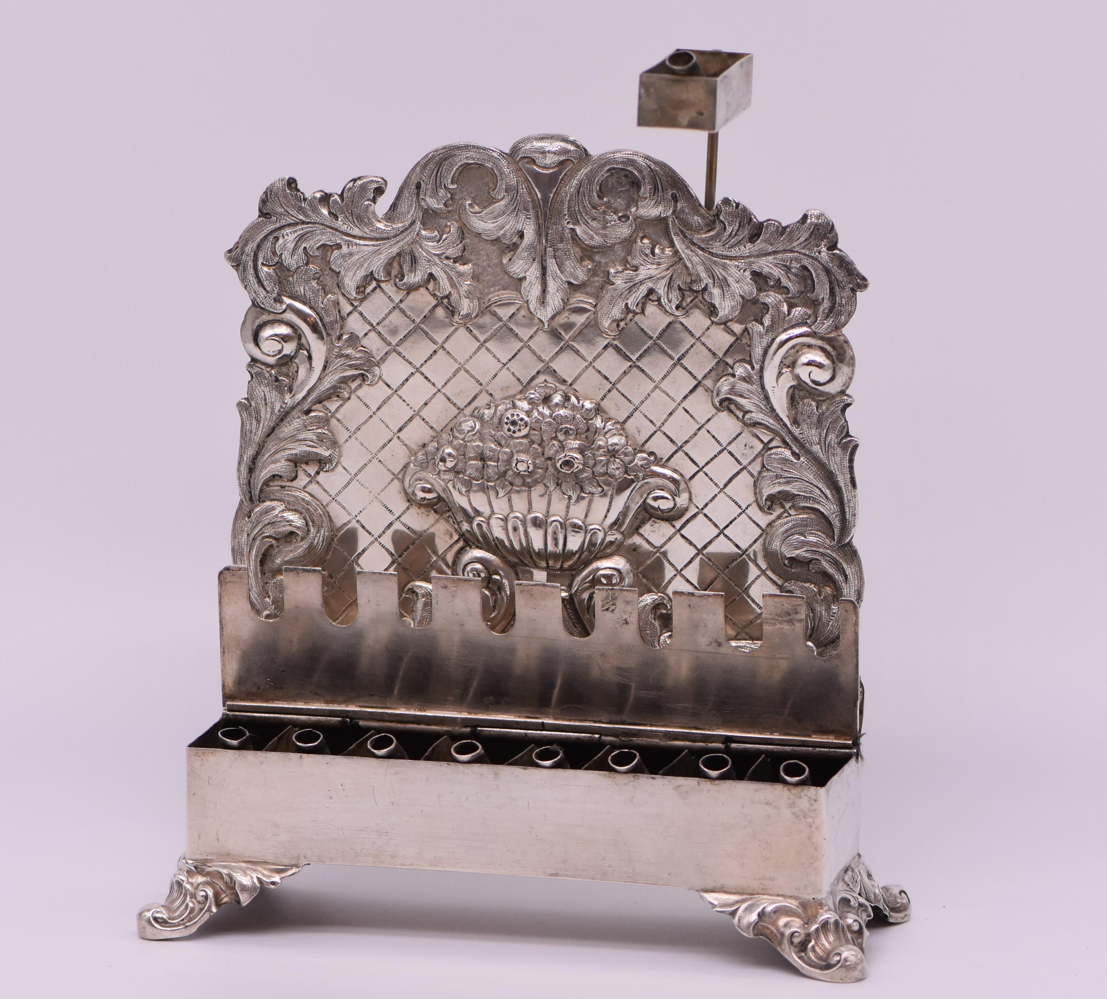 German silver Hanukkah lamp, marker's mark L.S. incuse, berlin, 1847-50. Seen in bench form with covered lamps, on shell and scroll feet. The backplate chased with an urn of flowers on trellis ground within embossed leaf border marked on the