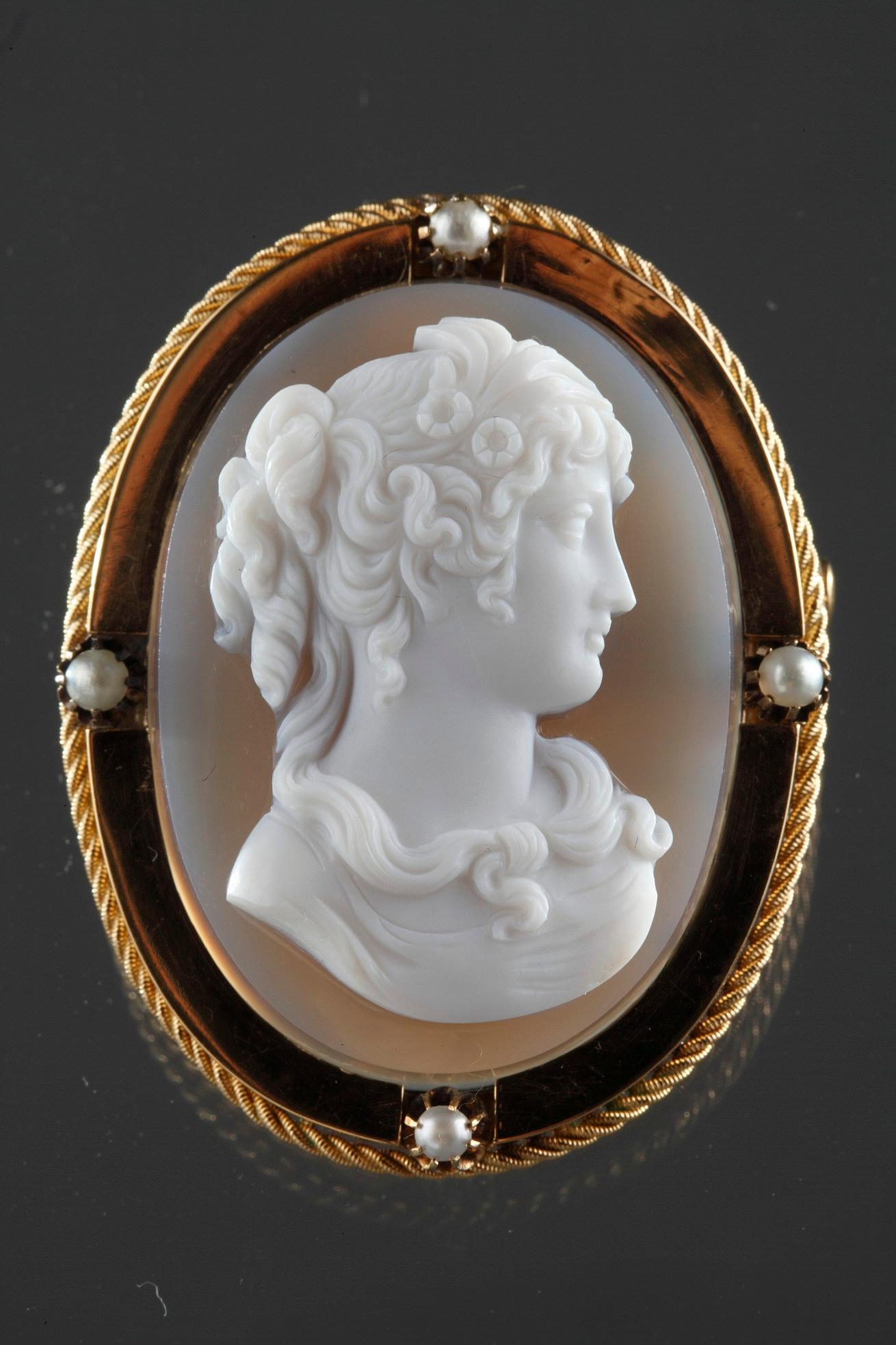 Cameo on blue-gray agate featuring a young woman looking toward the right. The artist intricately sculpted the white vein of the agate to bring the delicate profile to life. The young woman represented in bust has hair up. Strands of her hair fall