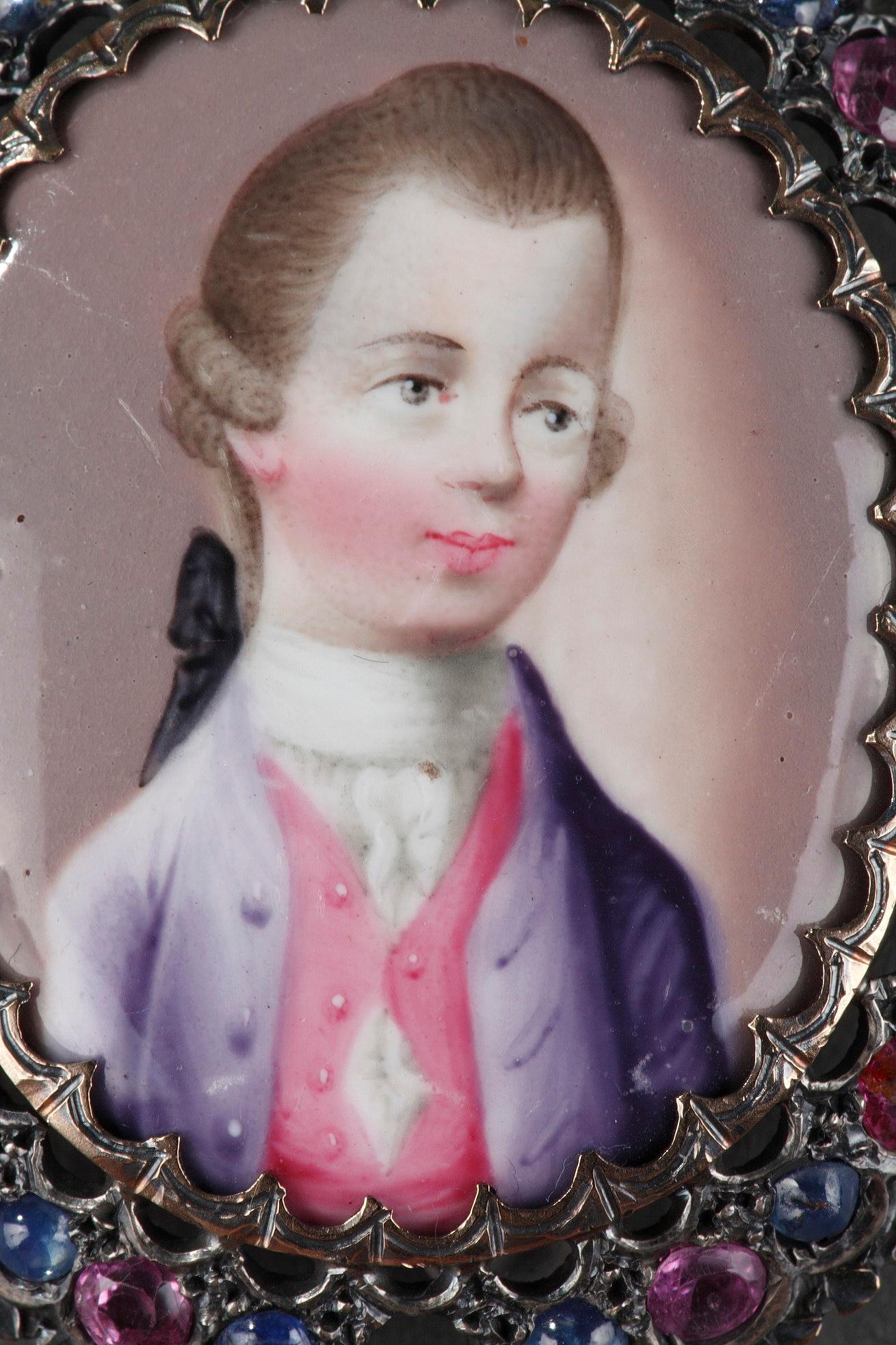 Eine ovale Miniatur auf Emaille mit dem Porträt eines eleganten Herrn. Sein Haar ist mit seitlichen Locken und einer großen Schleife frisiert, und er trägt Kleidung im Stil des 18. Jahrhunderts. Die Goldfassung ist abwechselnd mit Saphiren und