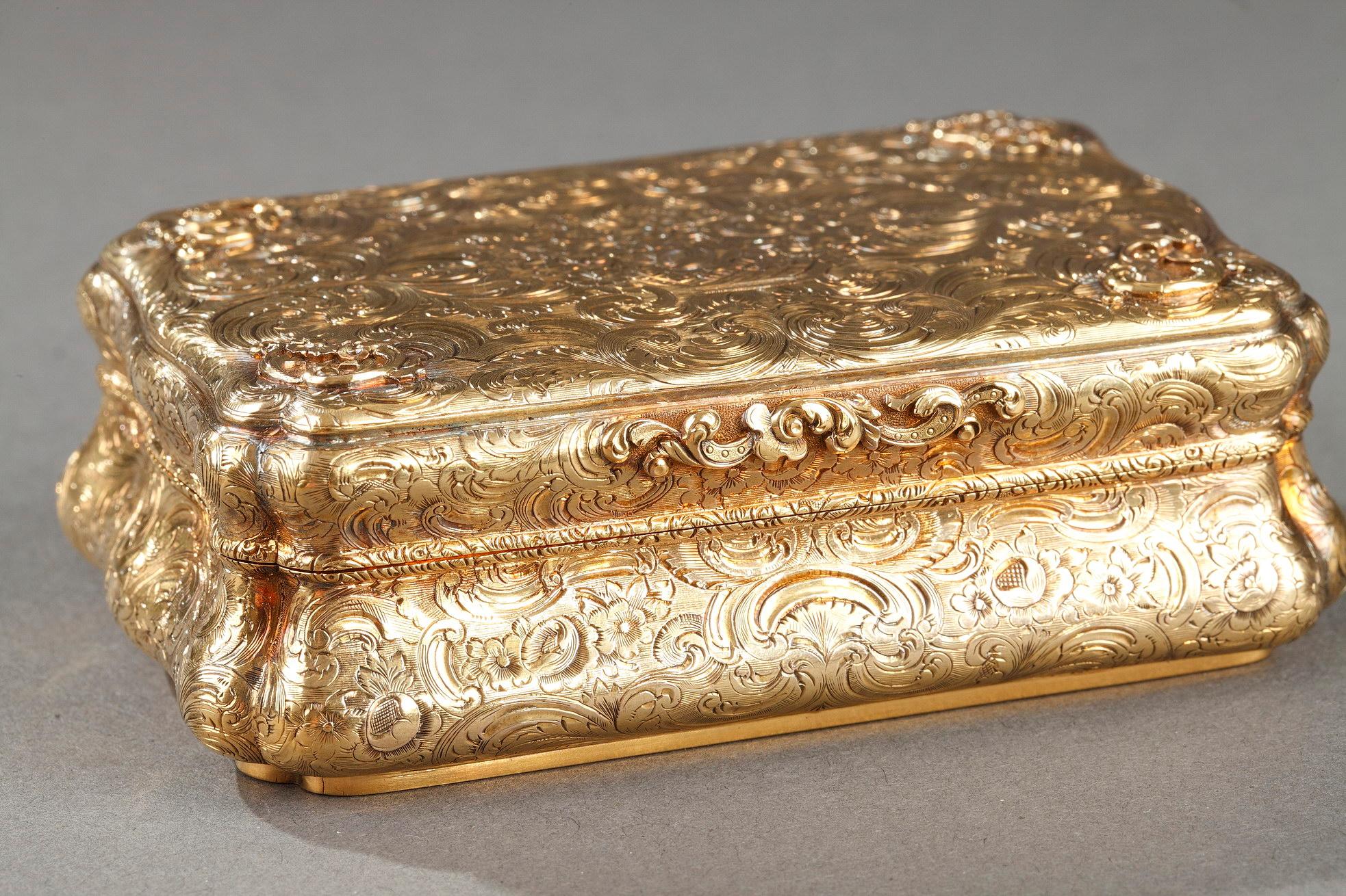 A cartouche-shaped gold box. The hinged lid, the base and the sides are chiseled with flowers and scrolls as well as rocaille patterns in slight relief. The lid has a chiseled bouquet of flower  in its center in a basket. On the back of the lid an