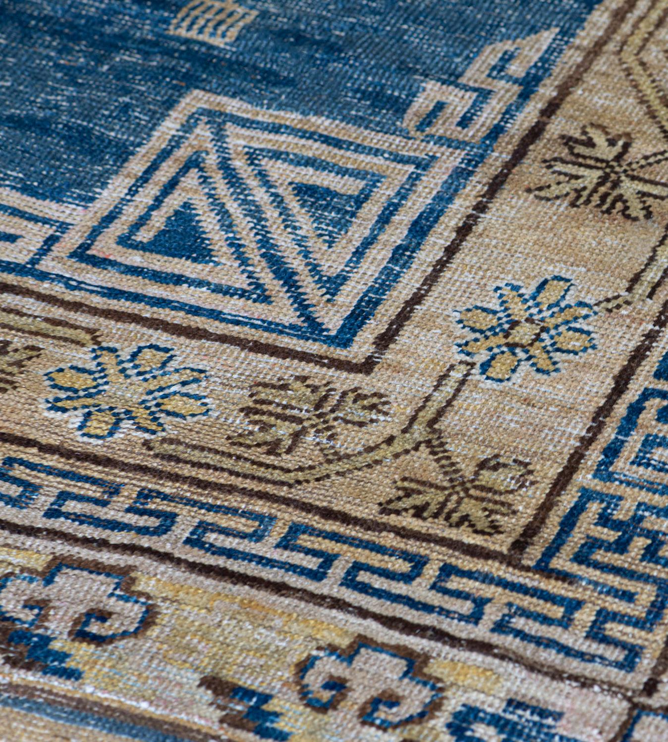 This traditional Khotan rug has a shaded blue field scattered with floral sprays and hooked lozenges together with floral vases and other auspicious motifs around a column of three beige roundels containing an outer band of rosettes and stellar
