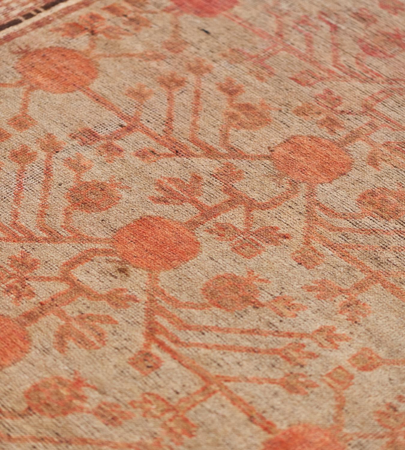 Ce tapis Khotan vintage présente un champ gris chaud avec un motif général de vigne de grenade rouge tomate ombrée émettant des têtes de fleurs angulaires, dans une bordure brun sable de têtes de fleurs liées abricot ombré, une bordure intérieure