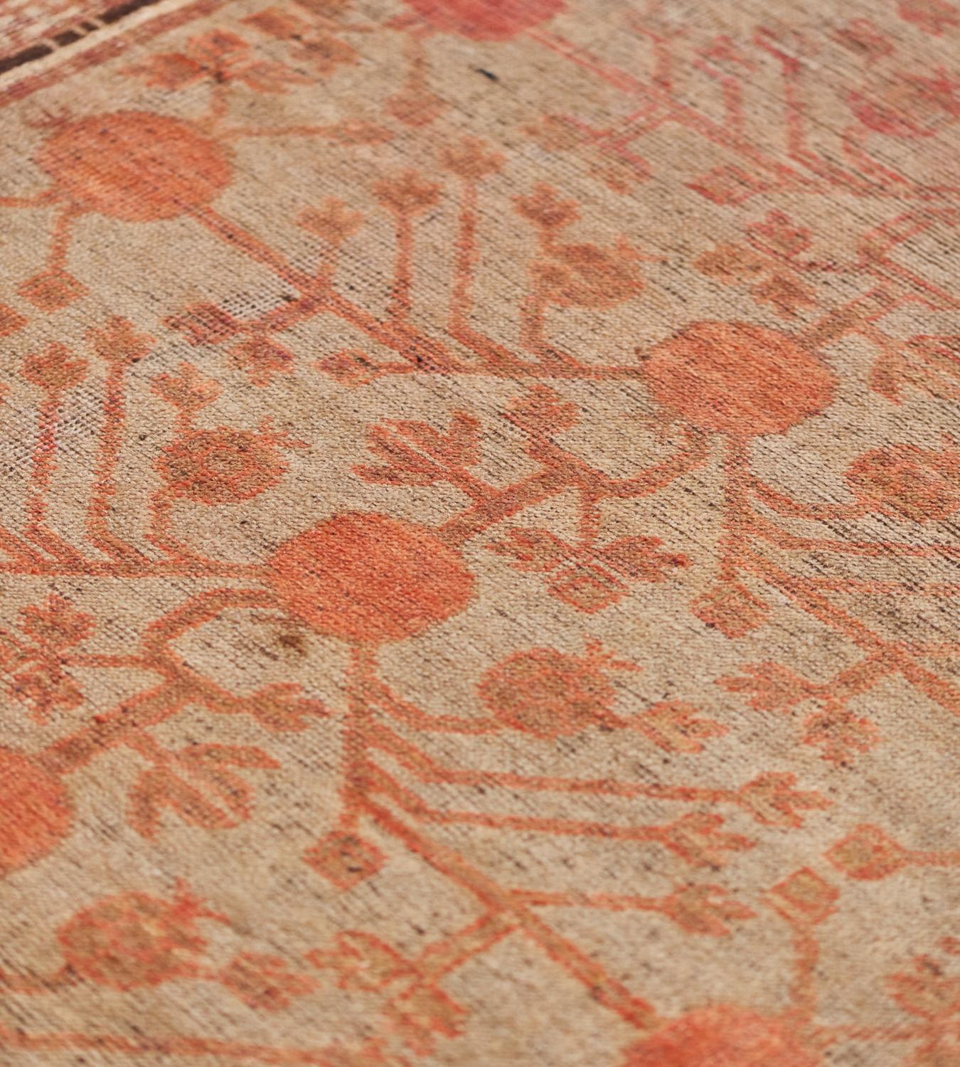 This vintage Khotan rug has a warm-gray field with an overall design of shaded tomato red pomegranate vine issuing angular flowerheads, in a sandy-brown border of shaded apricot linked flowerheads, an inner border containing polychrome square linked