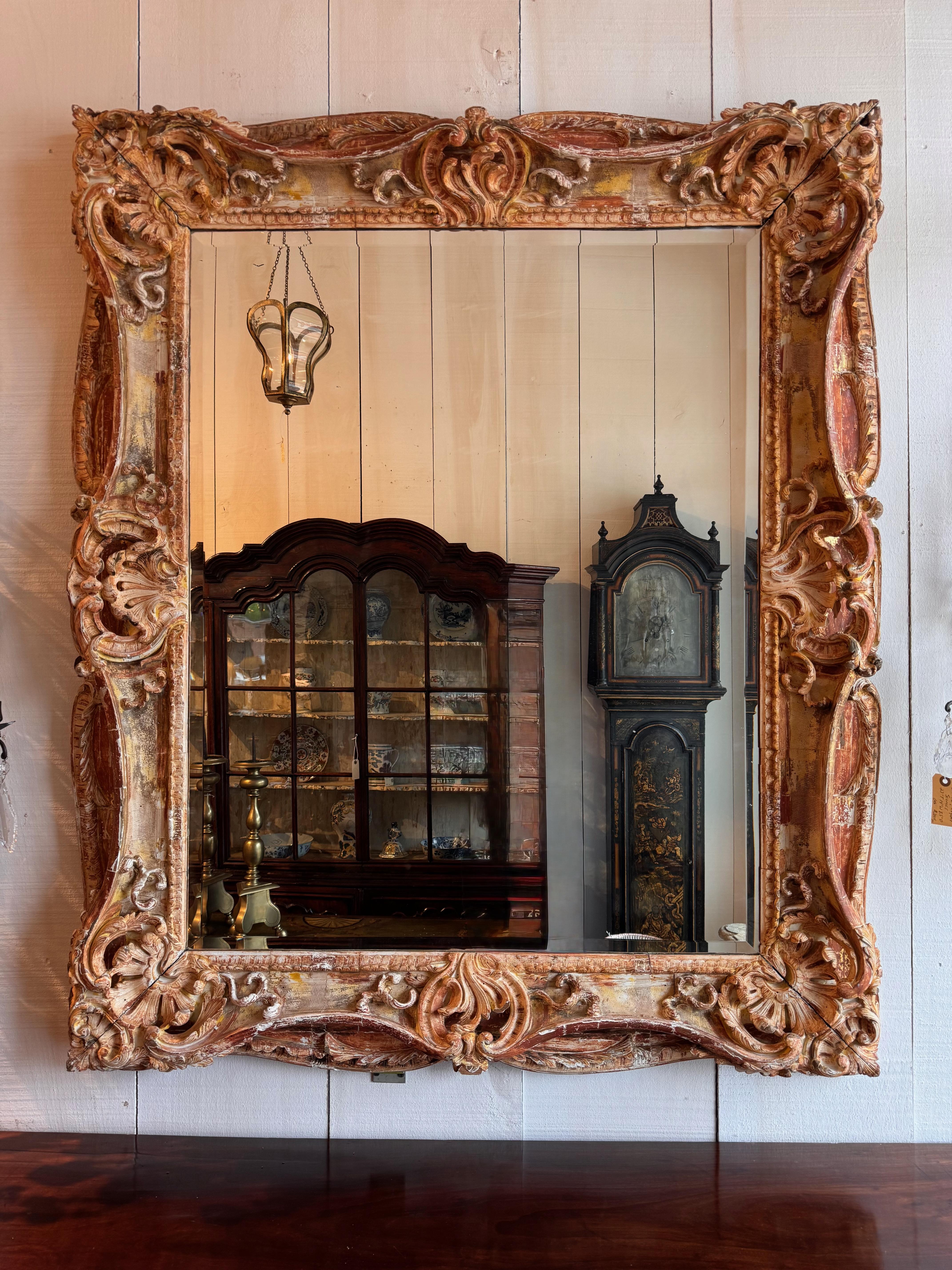 Ornately carved mirror in a bold Baroque or Rococo revival style, featuring a deep, scrolling frame with acanthus flourishes and shell motifs. The surface has a timeworn polychrome finish in warm terracotta, ivory, and gold tones, enhancing its