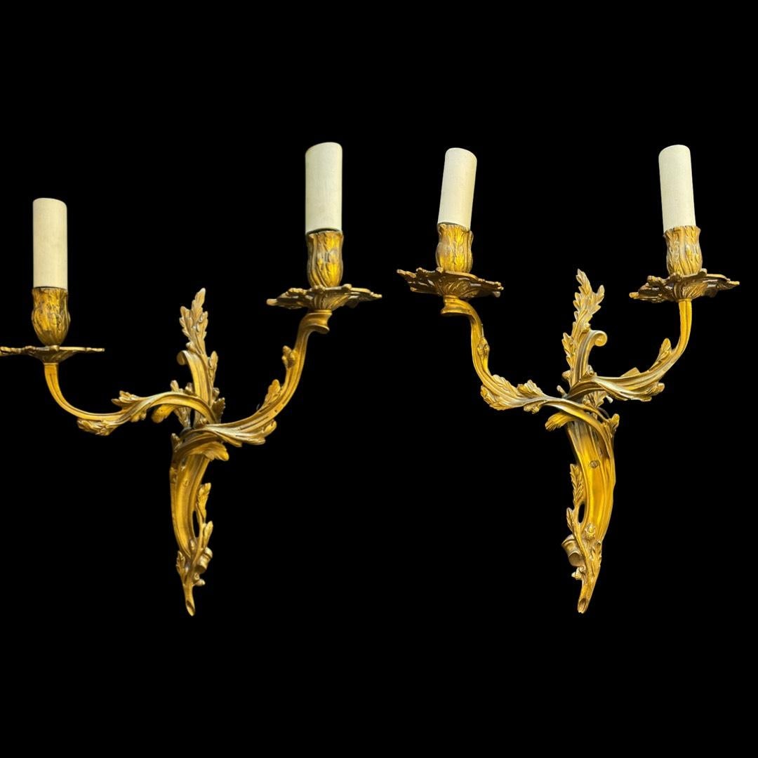 Ein charmantes Paar französischer Wandleuchter aus Messing im Stil Louis XV. Dieses entzückende Set besteht aus zweiarmigen Wandleuchtern, die jeweils mit Kerzenhaltern in Form von blühenden Blumen geschmückt sind. 

Inspiriert vom Rocaille-Stil