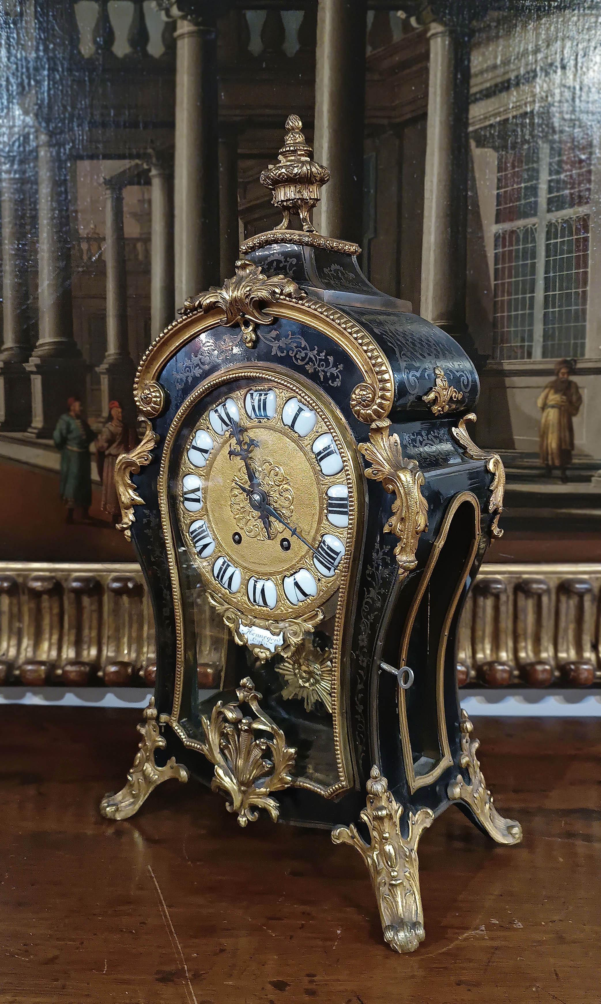 Elegant table clock with an arched case, crafted from hand-carved and ebonized wood, decorated with lost-wax cast and gilded bronze details. Further refinement is provided by the brass inlays on the wood, featuring plant and geometric motifs that