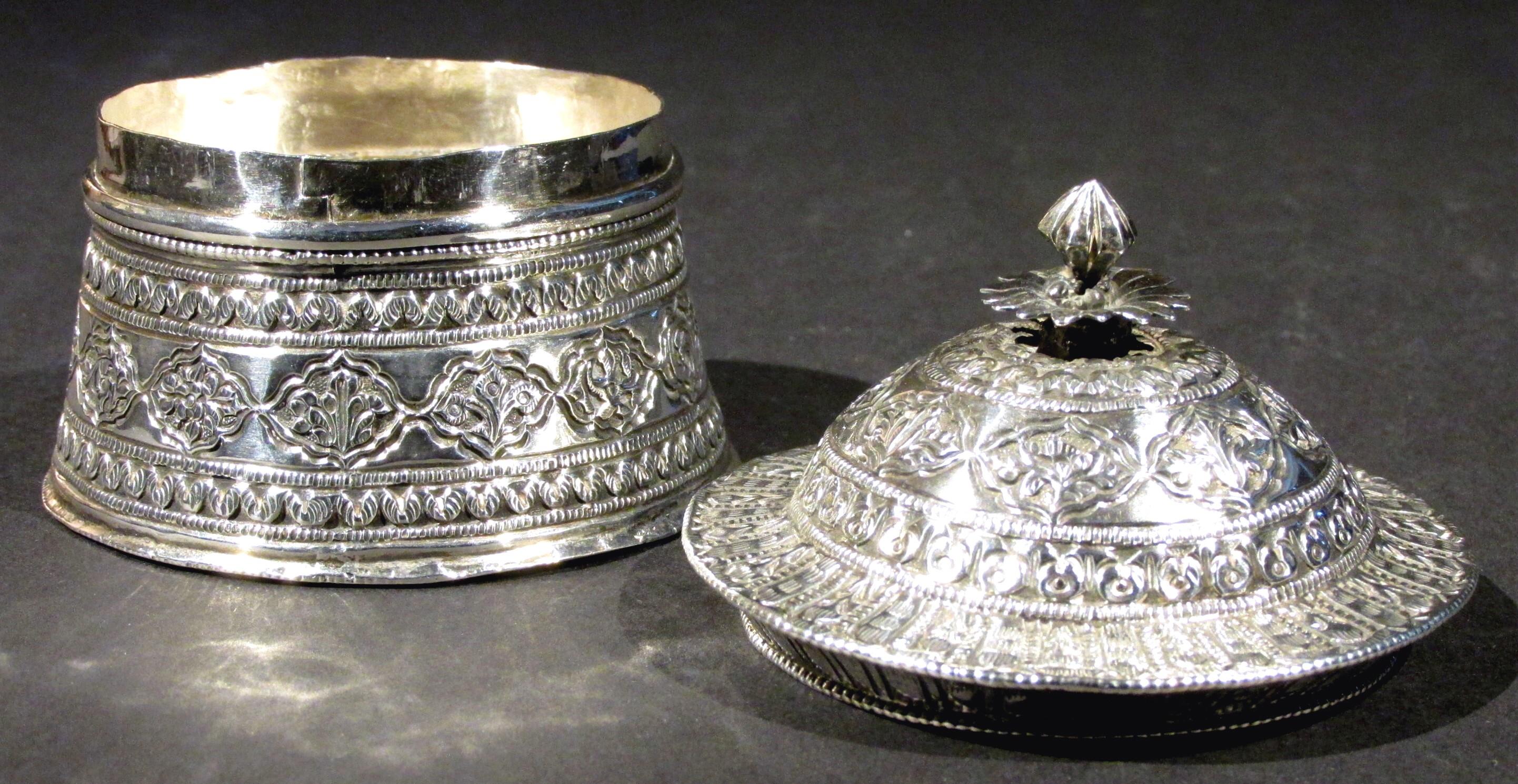 Intended to hold paan or betel, the seamed cylindrical body flaring downward towards the base, the domed lid showing a lotus-bud final over a flattened and canted rim, both decorated overall with repeating bands of hand embossed & chased geometric