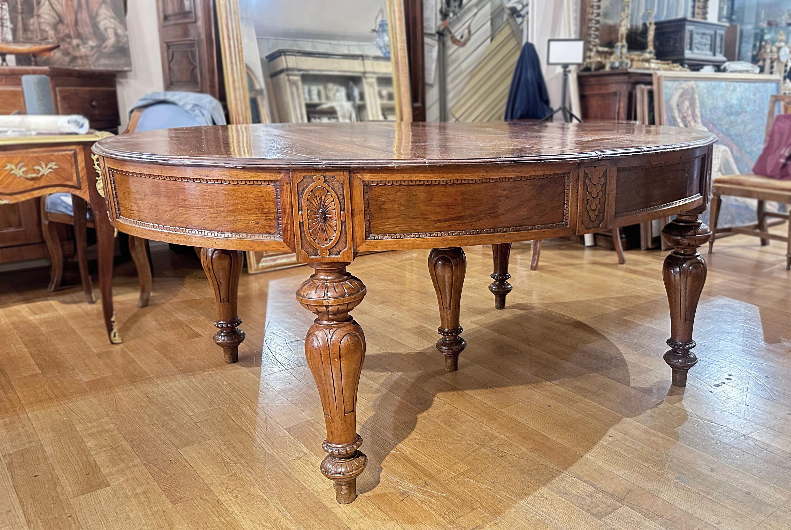 Elegant oval table in solid walnut, hand-carved, seating approximately eight people. The table features turned legs that support the under-table band, which is carved with geometric motifs and panels adorned with plant elements, enhancing its