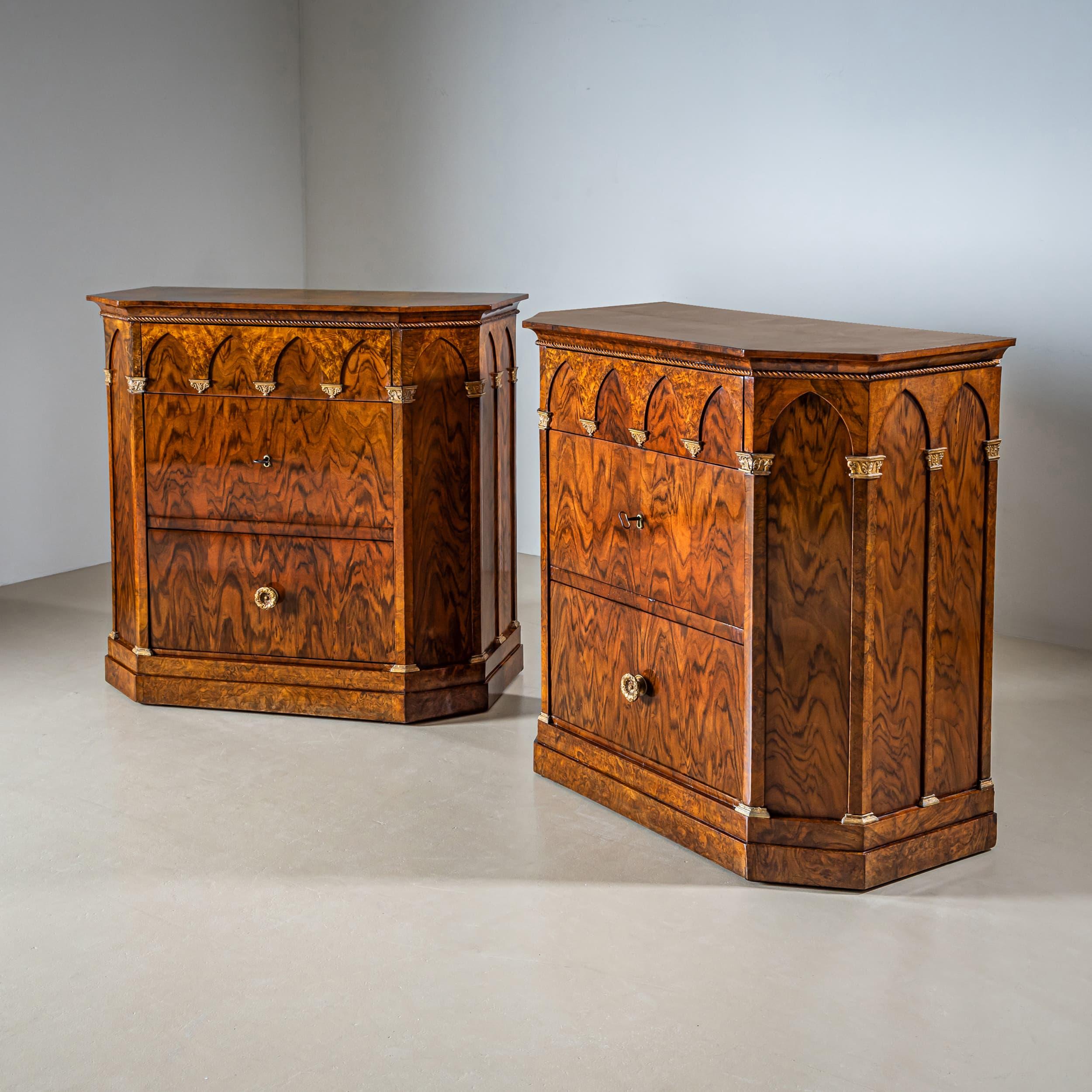 Elegante coppia di comodini realizzati in noce e impiallacciatura di radica, caratterizzati da angoli smussati, base dritta profilata e tre cassetti. Le decorazioni ad arco a sesto acuto in stile gotico con capitelli dorati ornano l'area sottostante