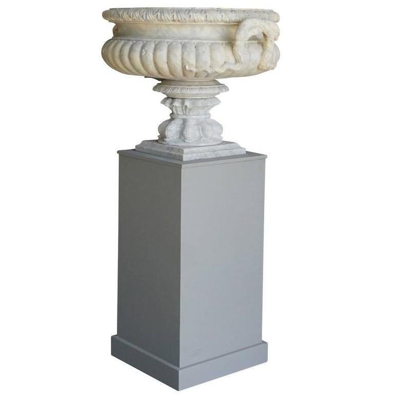 An antique French pair of shallow white Carrara marble shallow Tazza urns, the handles contain detailed and carved interlaced snakes, in good condition. The base of the vases are decorated with dolphins. The light grey wooden pedestals have been on