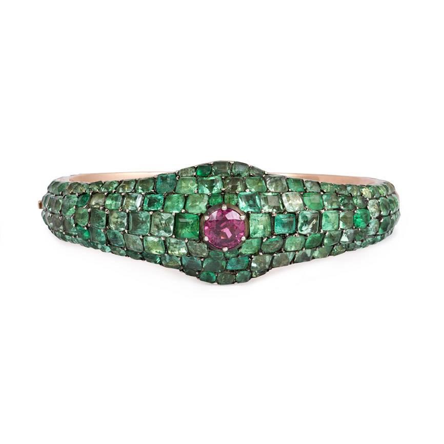 An antique hinged bangle bracelet of tapered design, pavé set all around with emeralds and centering on a pink sapphire, in silver and gold. 

Dimensions: 6.25 inches inner circumference, 7/8 inch at widest point