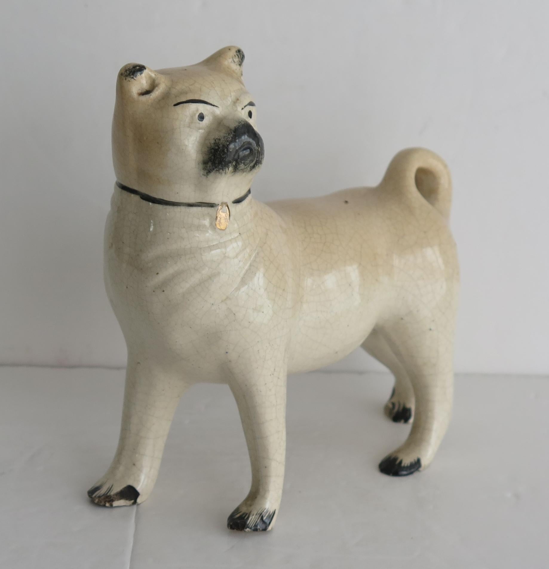 This is a good pottery figurine of a Pug Dog made by one of the Staffordshire potters, which we date to mid 19th Century early Victorian England.

The dog figure is well modelled then hand painted with a gilded name tag to the collar.

There are