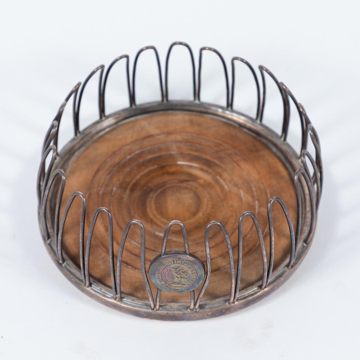 Mid-19th-century Sheffield silver champagne coaster with carved wooden base. The looped wire pickets create a supportive buttress for a bottle or flask, and feature a small commemorative or heraldic disc accent, inscribed with a carving of two birds