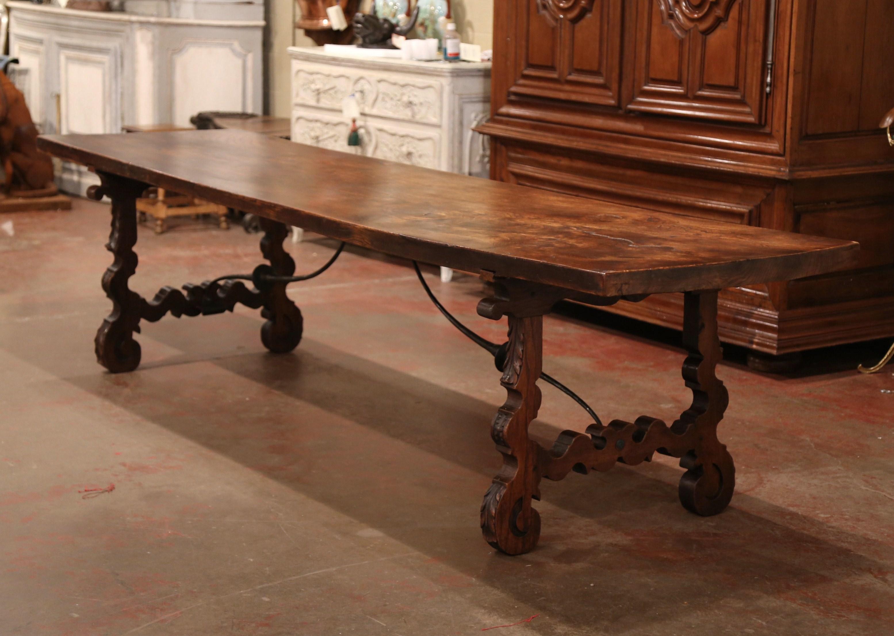 This long antique fruitwood table was crafted in Spain, circa 1850, the elegant table stands on two carved pedestal legs decorated with acanthus leaves and connected with a forged black iron stretcher for stability and structure. The thick top is