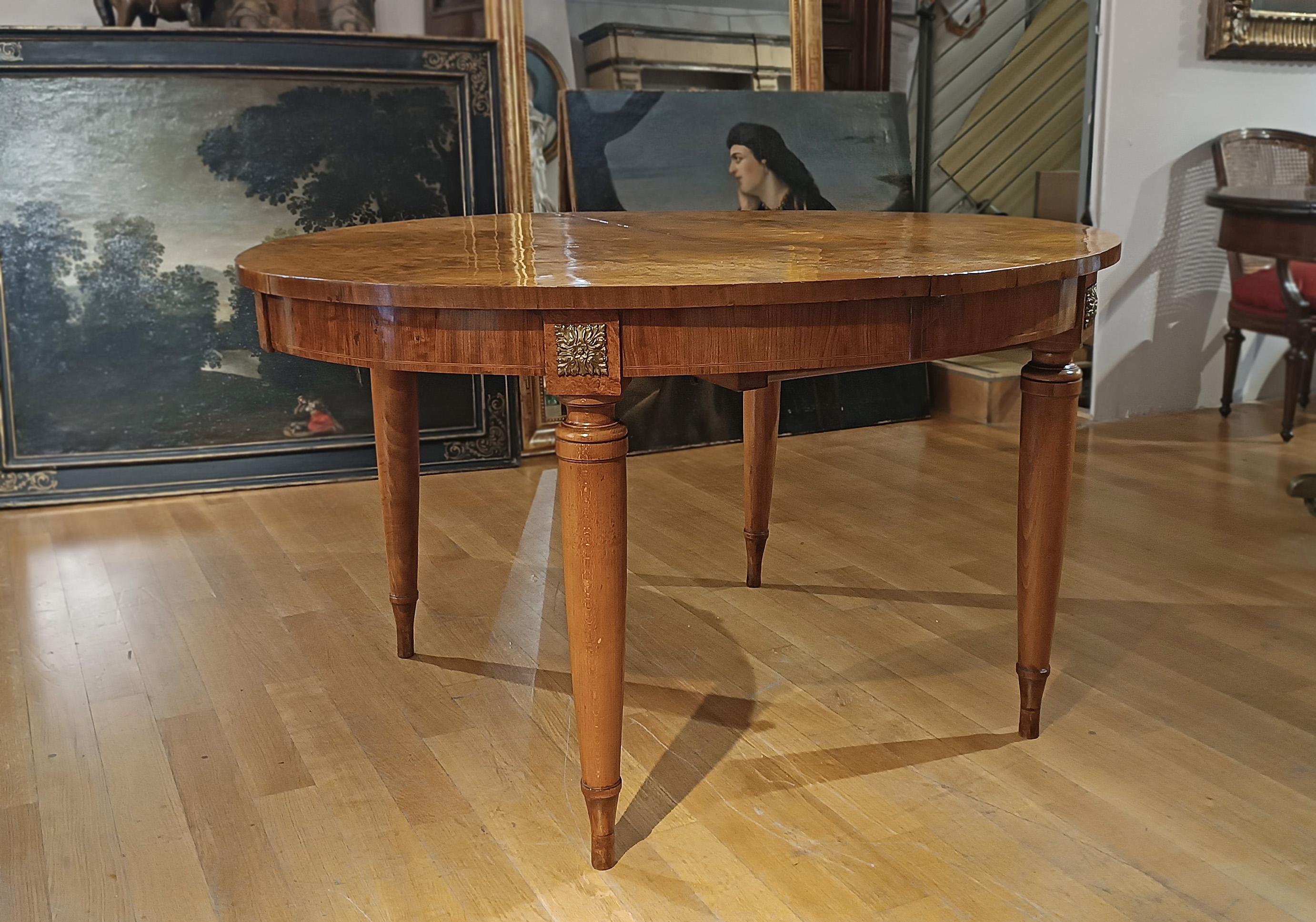 Refinada mesa de comedor ovalada de tuya tallada a mano. La mesa tiene cuatro patas rectas con pies puntiagudos y una banda bajo el tablero decorada con detalles florales en bronce a la cera perdida. La encimera presenta incrustaciones geométricas