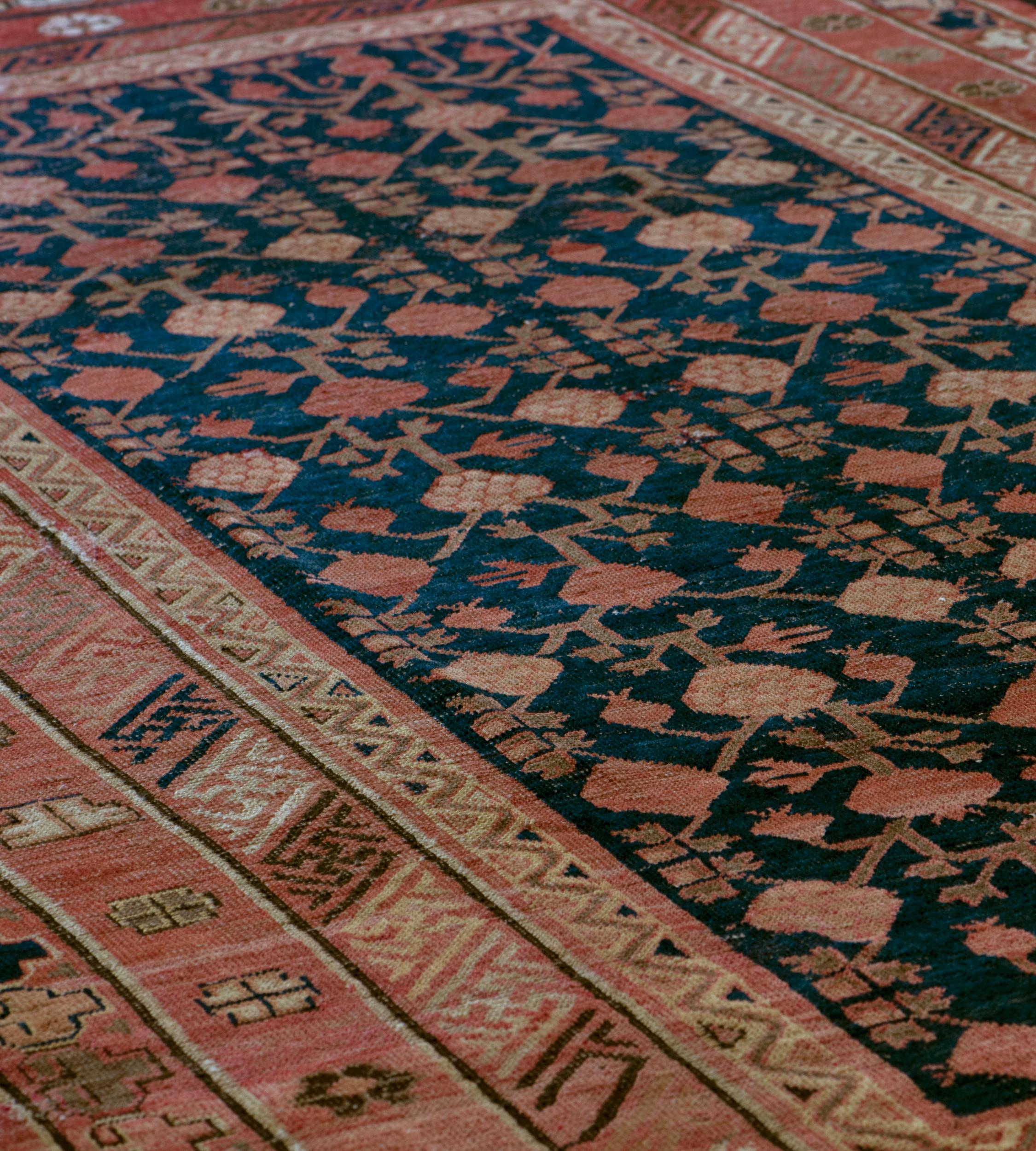 This antique Khotan rug has a shaded indigo-blue field with an overall design of angular pomegranate and leafy motif vine, in triple striped borders of shaded soft terracotta-red spaced flowerheads, diagonal key-pattern and stylised cloud motifs,