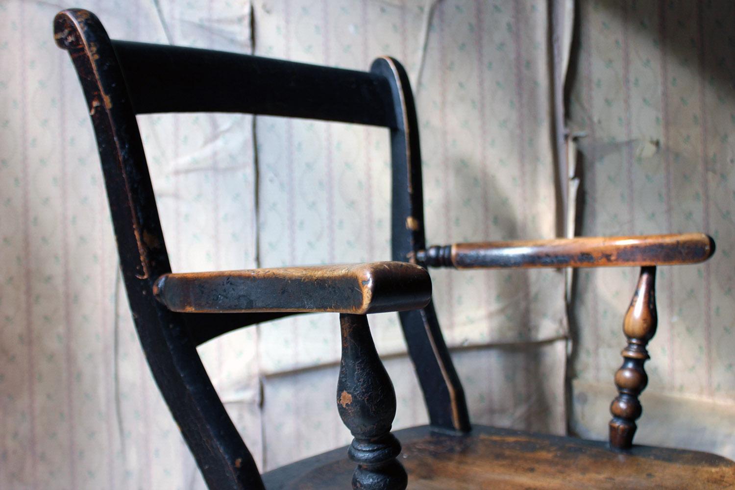 The beautifully time-worn farmhouse black painted lath backed elm kitchen armchair, of good quality construction, having an elm slab saddle seat to turned arm supports and splayed legs, the whole surviving from the third quarter of the nineteenth