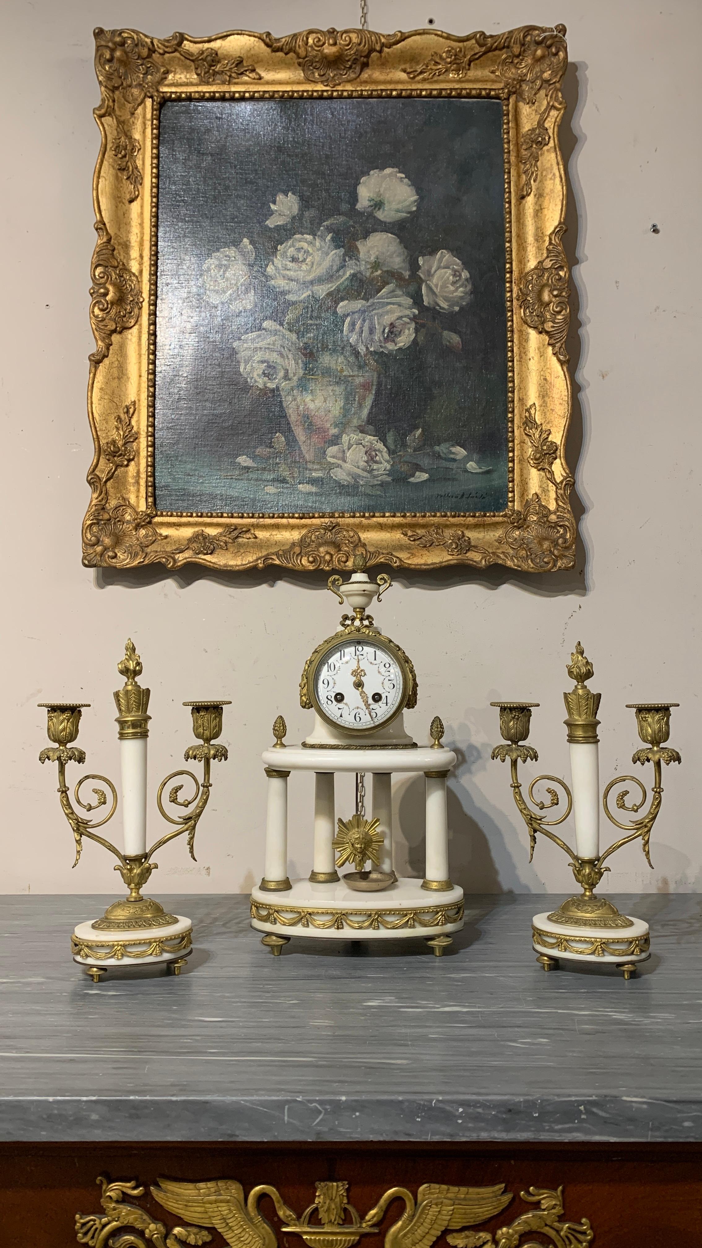 Triptychon bestehend aus einer Uhr und zwei Kerzenständern aus weißem Marmor mit vergoldeter Bronze. Die Uhr hat einen Wochenaufzug, mit Stunden- und Halbstundenschlag, die Scheibe kann geöffnet werden. Außerdem ist er mit einer