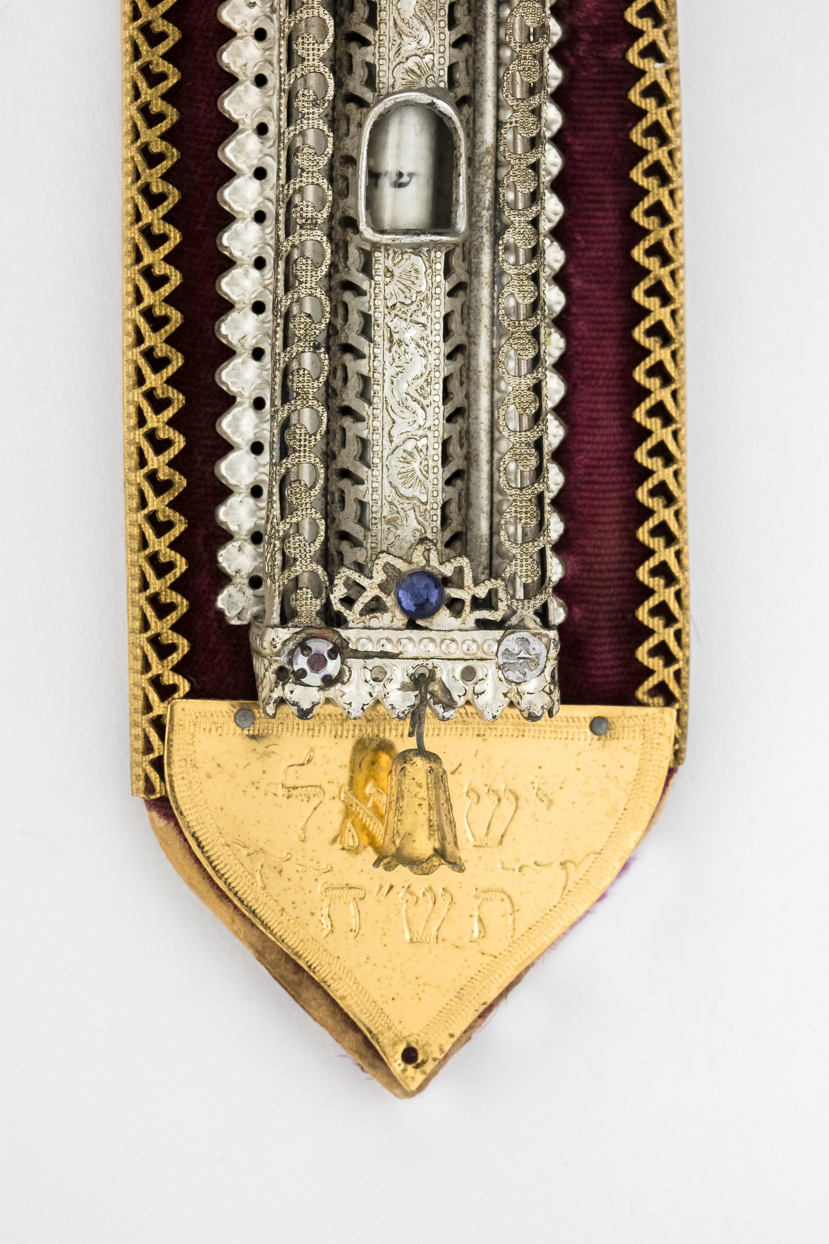 Early Israeli Mezuzah on velvet mounting with ornate silvered body, and with gilded top and base. Decorated with colored glass and with gilded bell hanging from the lower part of the Mezuzah. 
Engraved in Hebrew “Israel 1948” at base.
The Mezuzah is