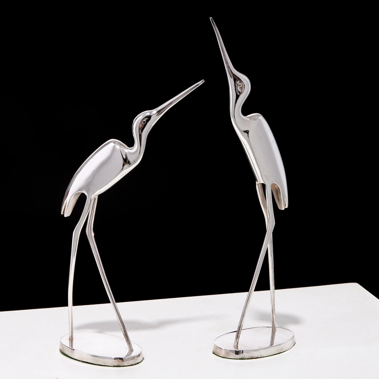 Un par de esculturas cromadas, elegantes y modernistas, que representan grullas al estilo del dúo de diseñadores Curtis Jeré. Estas aves son elegantemente alargadas, con largas patas y cuellos, y están colocadas para resaltar sus gráciles posturas,