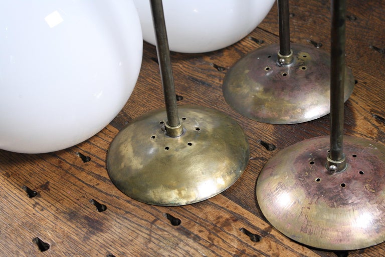 A run of 8 large globe opaline glass pendants with their original brass monk cap galleries, brass stems and spun brass ceilings roses.

All metal sections have age related oxidization, all glass is in great condition. 

Removed from Abbots