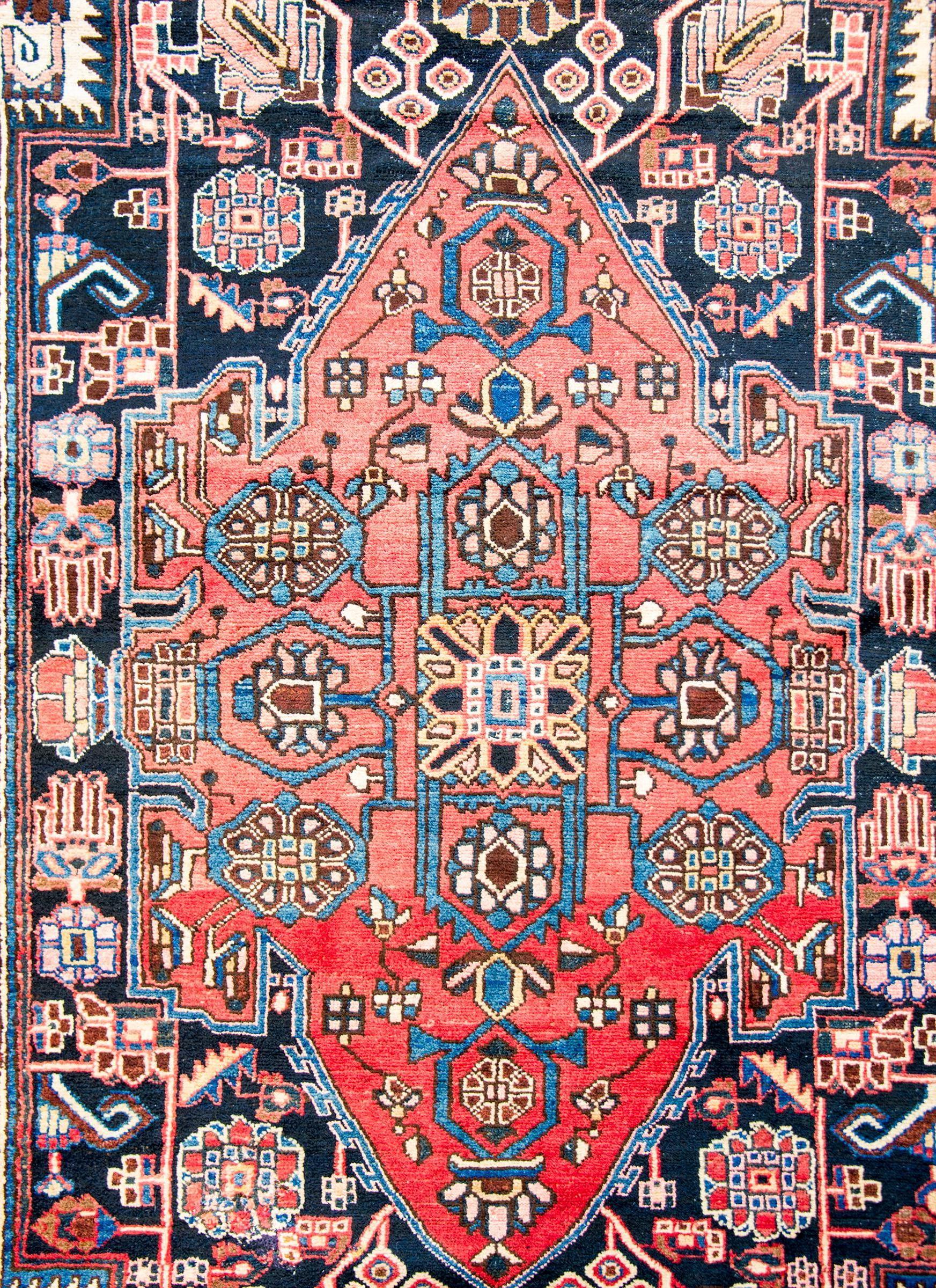 A mid-20th century Persian Mazlaghan rug with a fantastic mirrored tribal pattern with a large abrash crimson medallion with floral and scrolling vine pattern, on an indigo background with similarly styled flowers. The border is thin, with a central