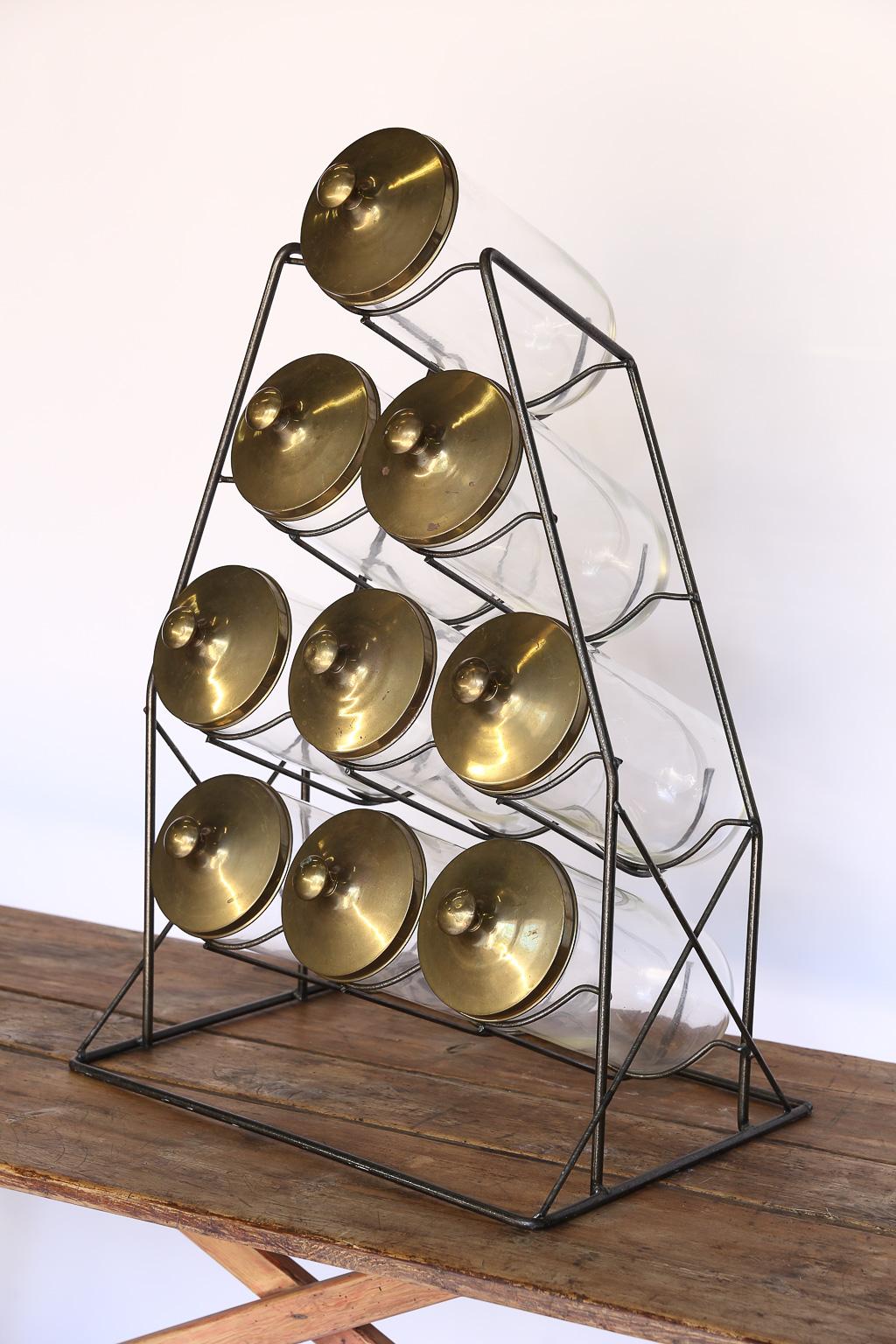 Found in England, this mid-20th century apothecary set is complete with nine lidded glass jars and iron Stand. The brass-plated metal lids measure 6.25 inches in diameter and 3 inches high, the glass jars measure 6 inches in diameter and 13.5 inches