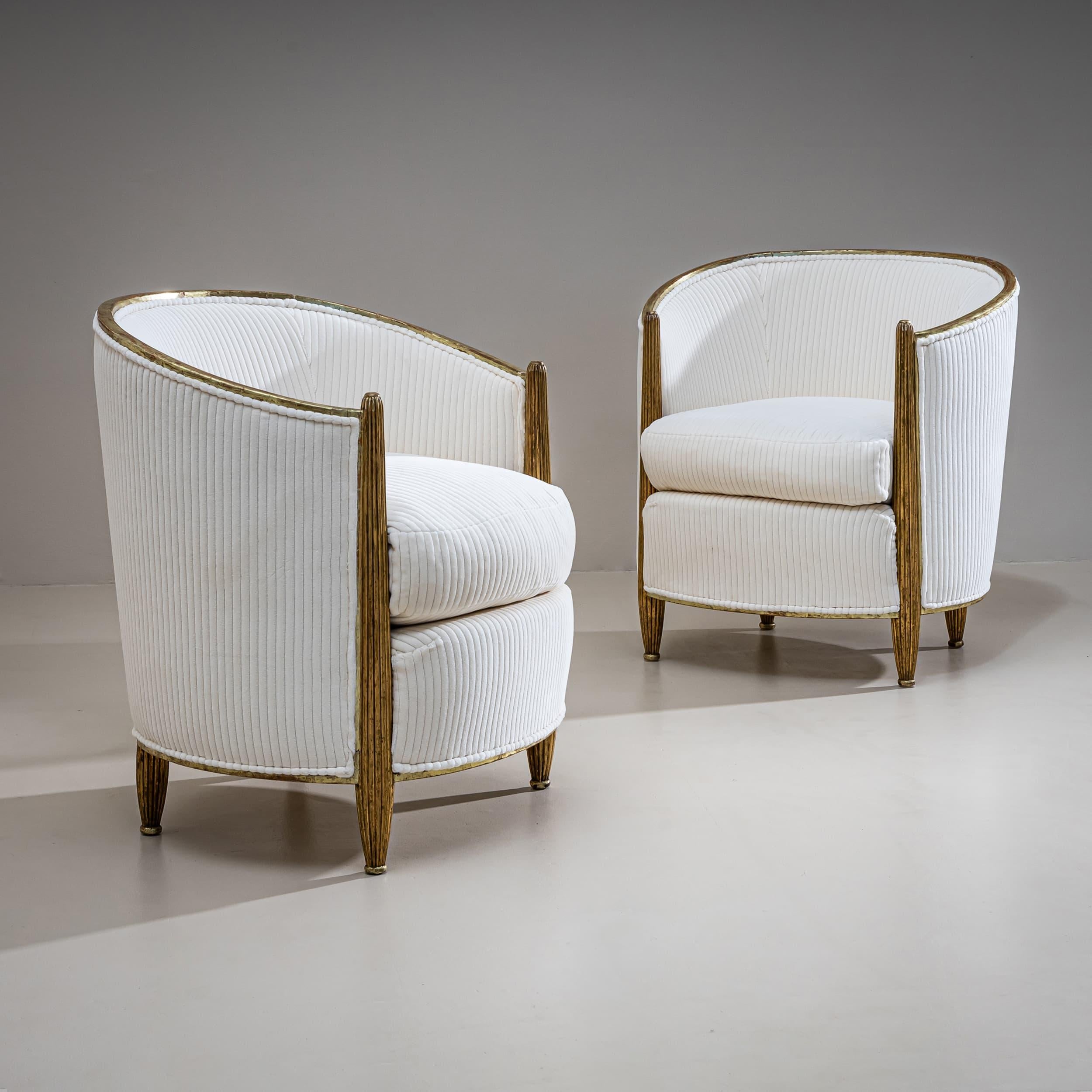 Pair of Art Deco armchairs featuring tapered, fluted legs and rounded backrests. The chairs are finished with a gold patina. The ribbed upholstery in cream white is newly fitted.