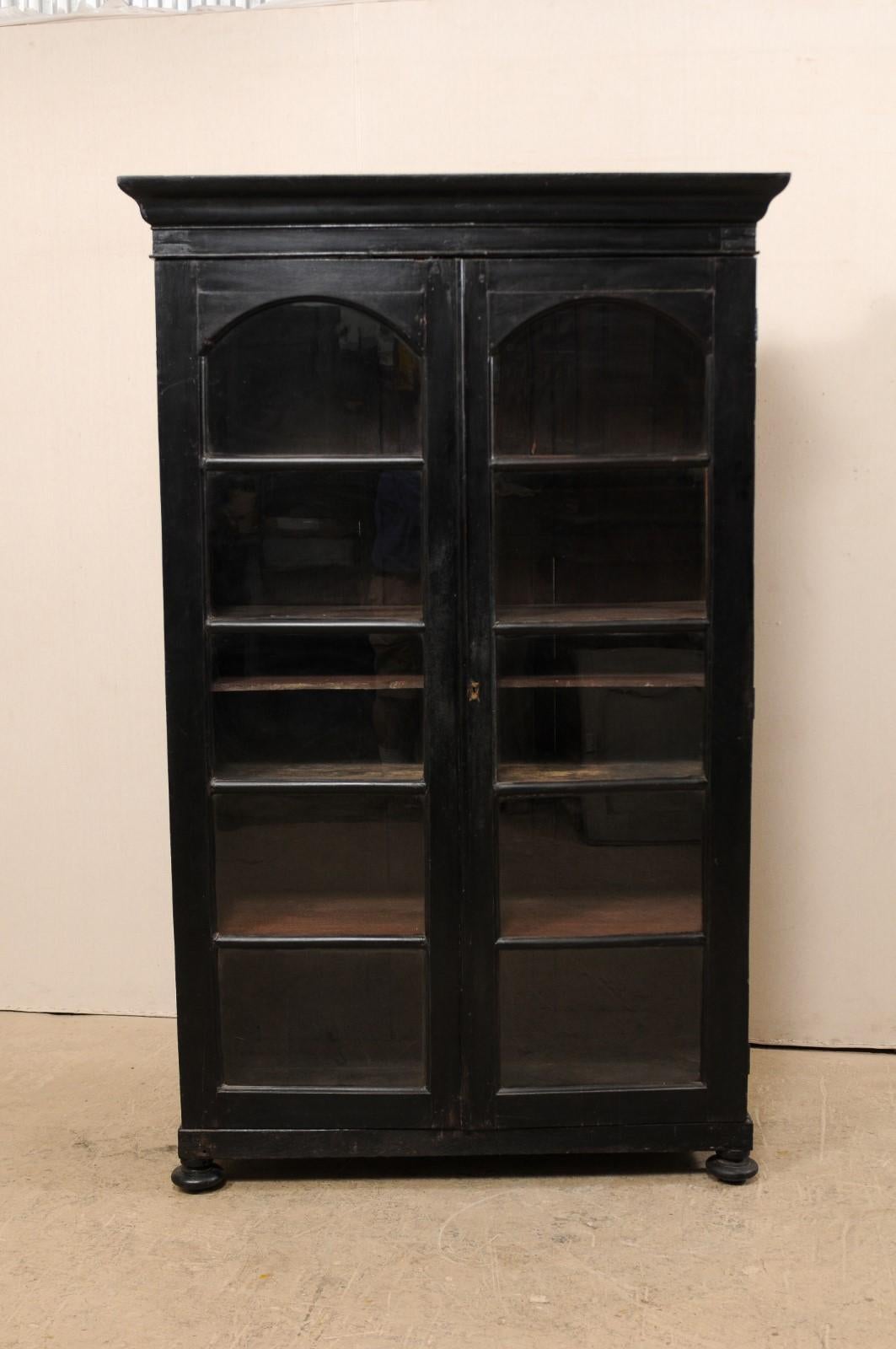 A tall British Colonial storage cabinet with glass doors from the mid-20th century. This vintage display cabinet features a crown-mold cornice atop a case with pair of arched panel front doors, set with glass. The skirt and sides of the cabinet