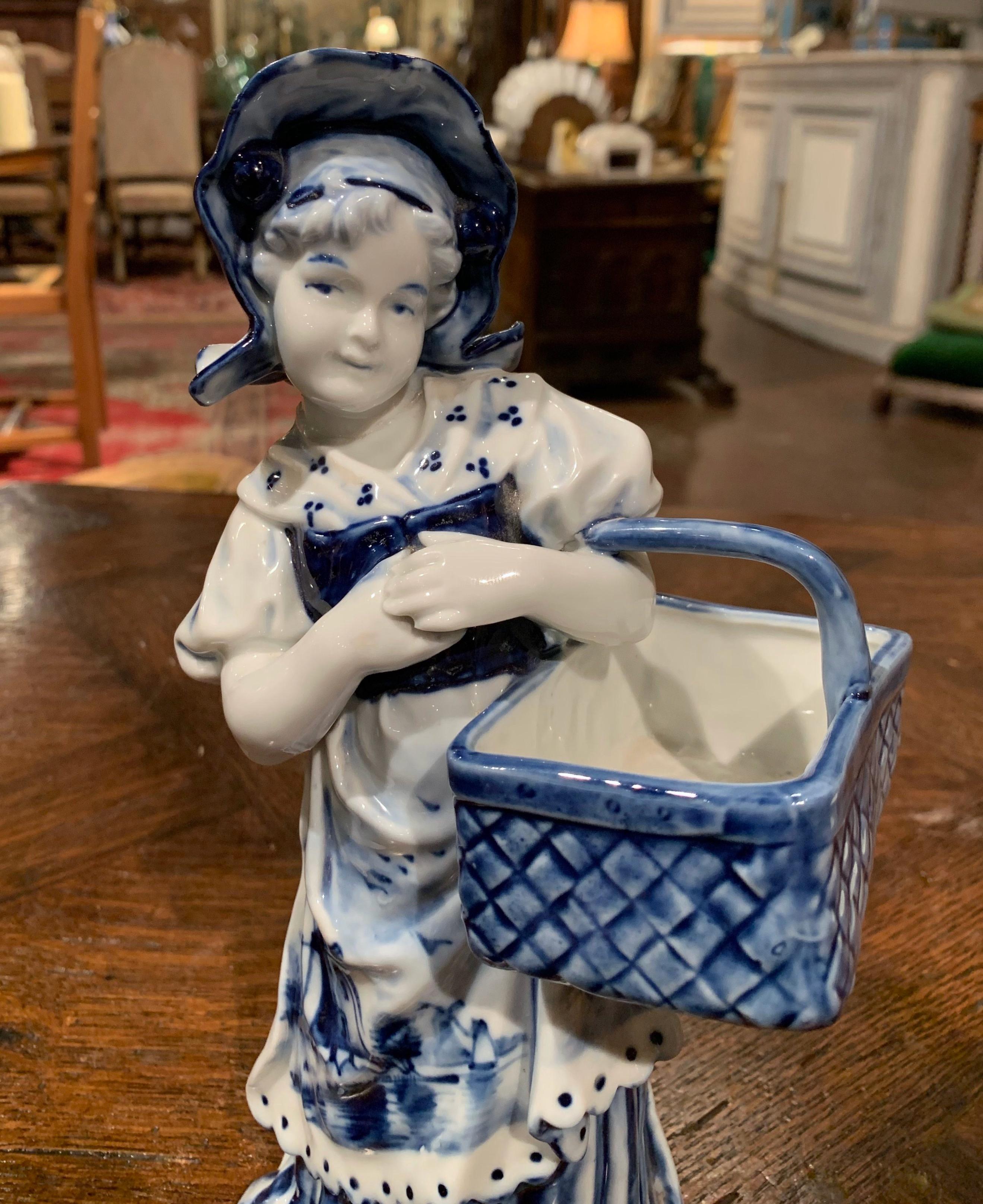 Crafted in Holland, circa 1950, the porcelain sculpture features a young beauty carrying a basket; the sculpture is hand painted in the traditional delft blue and white colors with floral and sailboat motifs on her dress. The figure, stamped on the