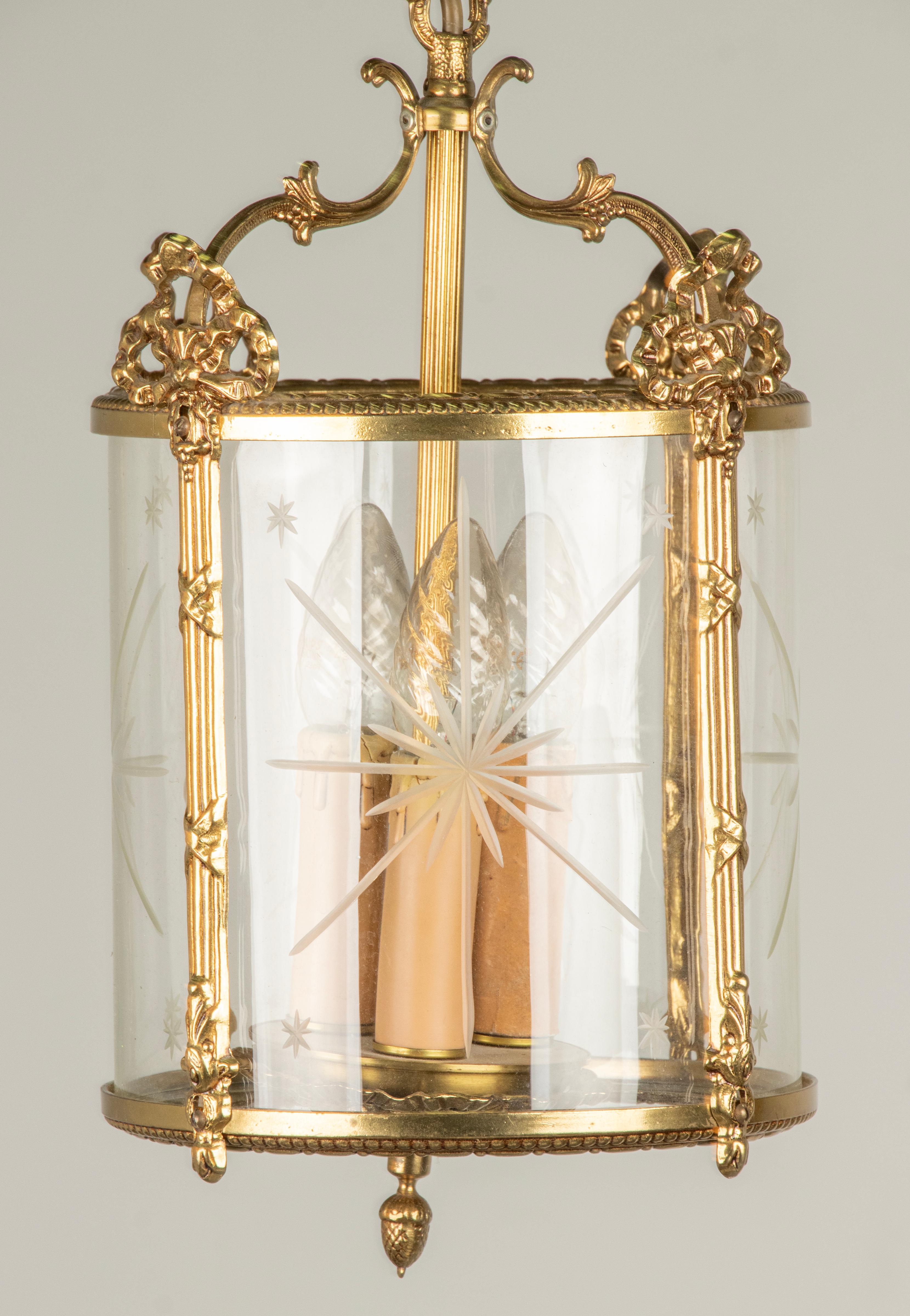 Lampe-lanterne en laiton de style Louis XVI. Décorés avec des rubans et des nœuds. A l'intérieur, un abat-jour en verre avec un motif d'étoiles taillées. A l'intérieur, il y a de la place pour 3 douilles pour des ampoules E14. 
Fabriqué en Belgique,